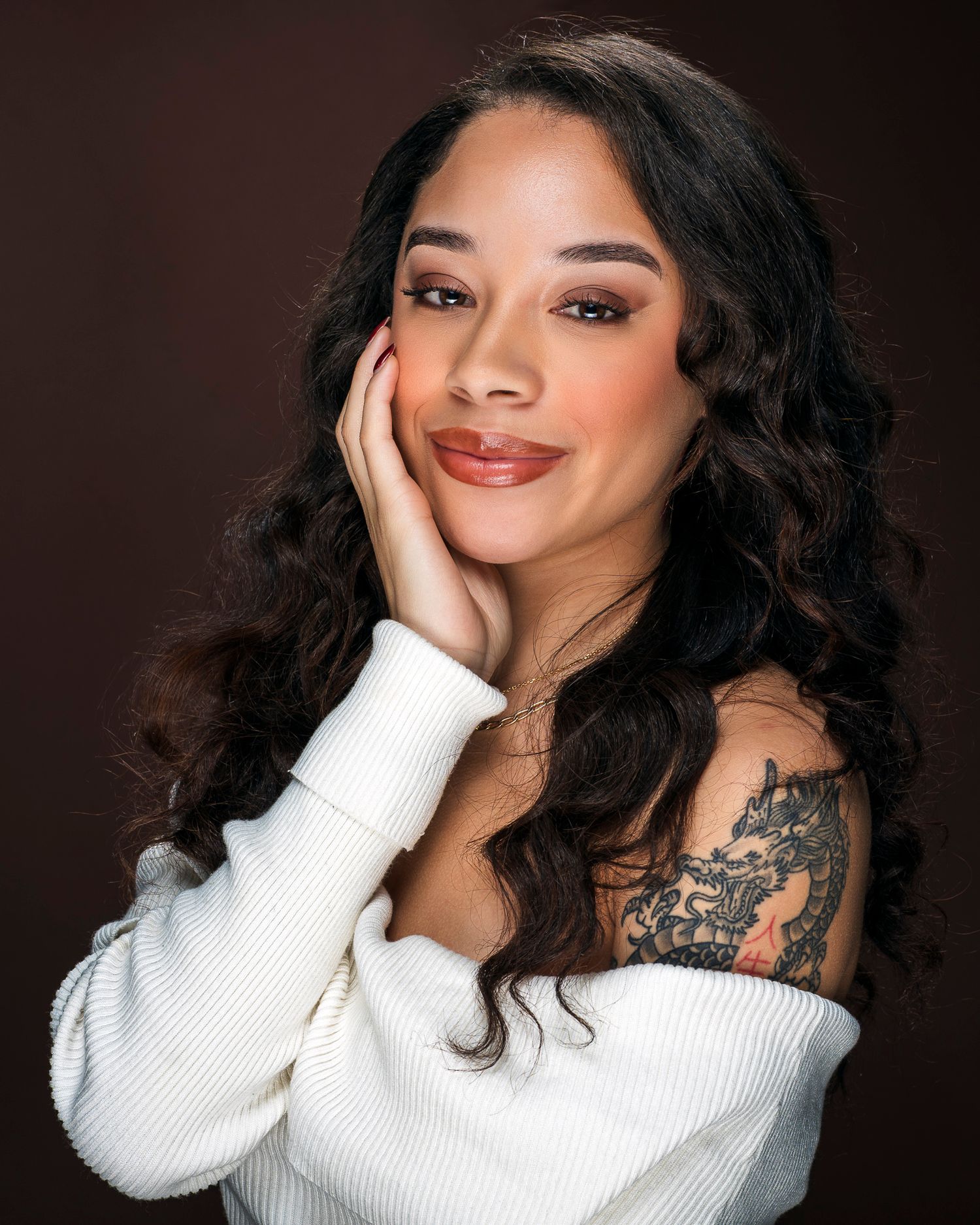 Beautiful woman with dragon shoulder tattoo and curly hair smiling in off-shoulder white sweater.