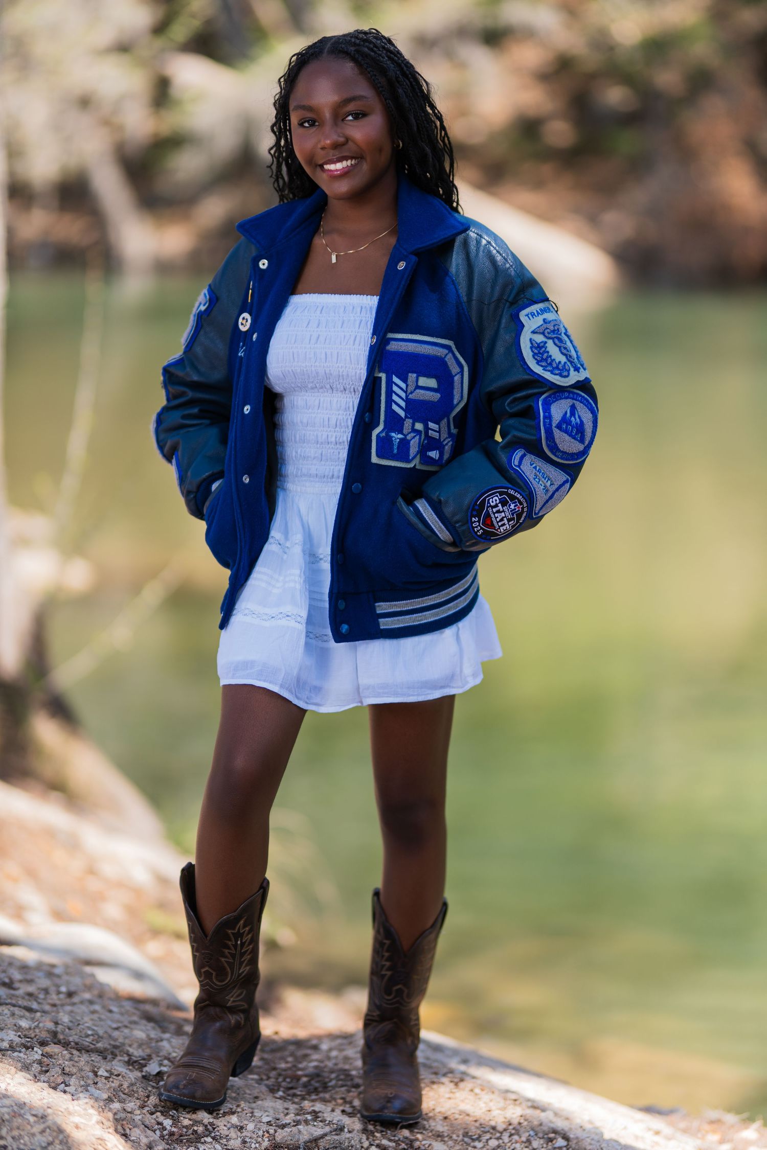 Young woman smiling in blue varsity jacket, white dress, and cowboy boots outdoors near water.