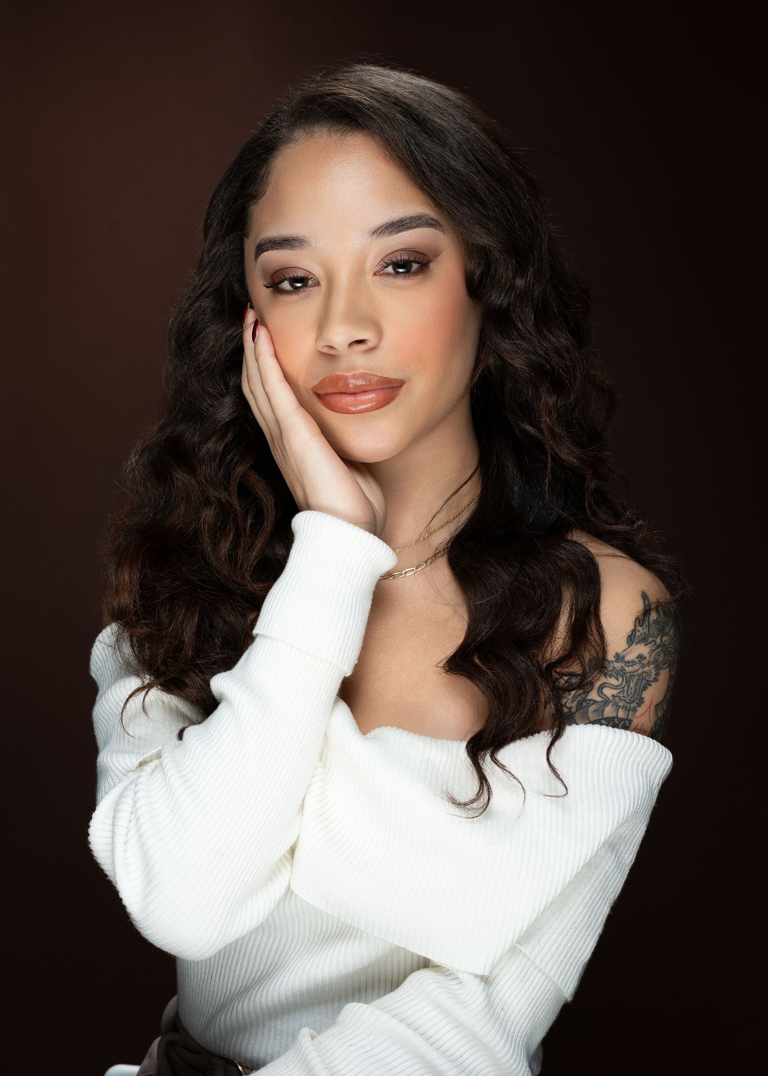 Young woman with long curly dark hair wearing white off-shoulder sweater, posing with hand on cheek against dark background.