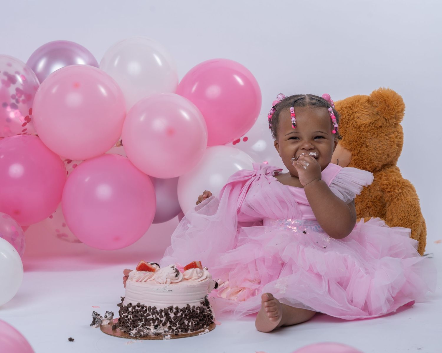 A young girl in a pink tutu dress celebrates her birthday with a cake, teddy bear, and pink balloons.