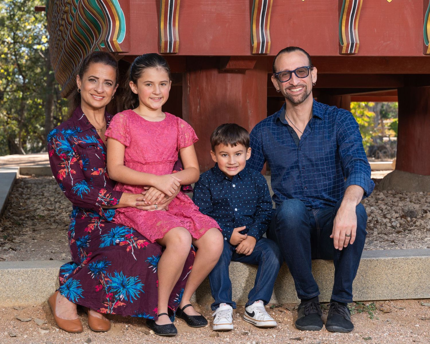 Happy family of four smiles together outdoors near colorful decorative structure in park setting.