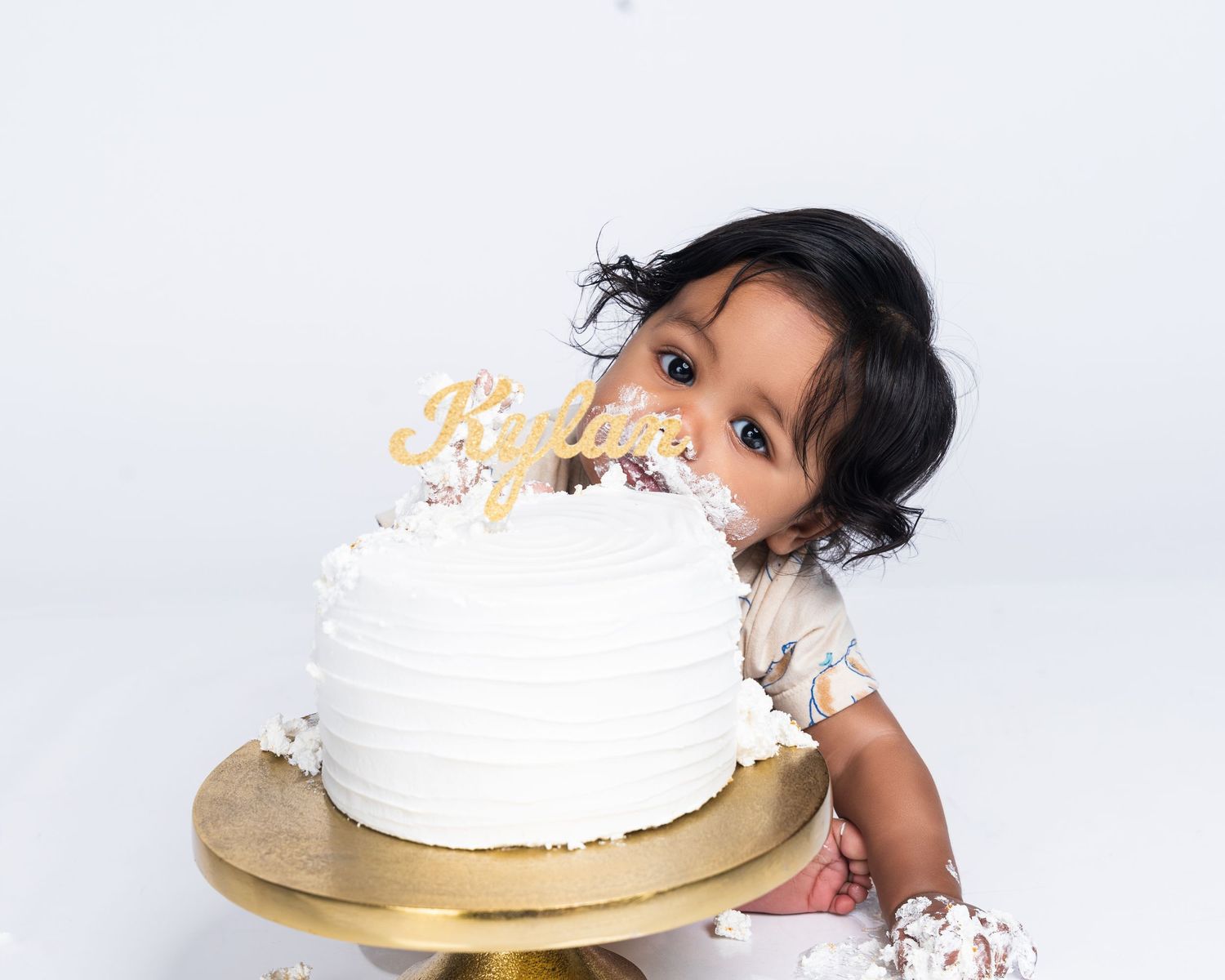 A baby girl peeks over a white frosted birthday cake with a gold name topper on a wooden cake stand.