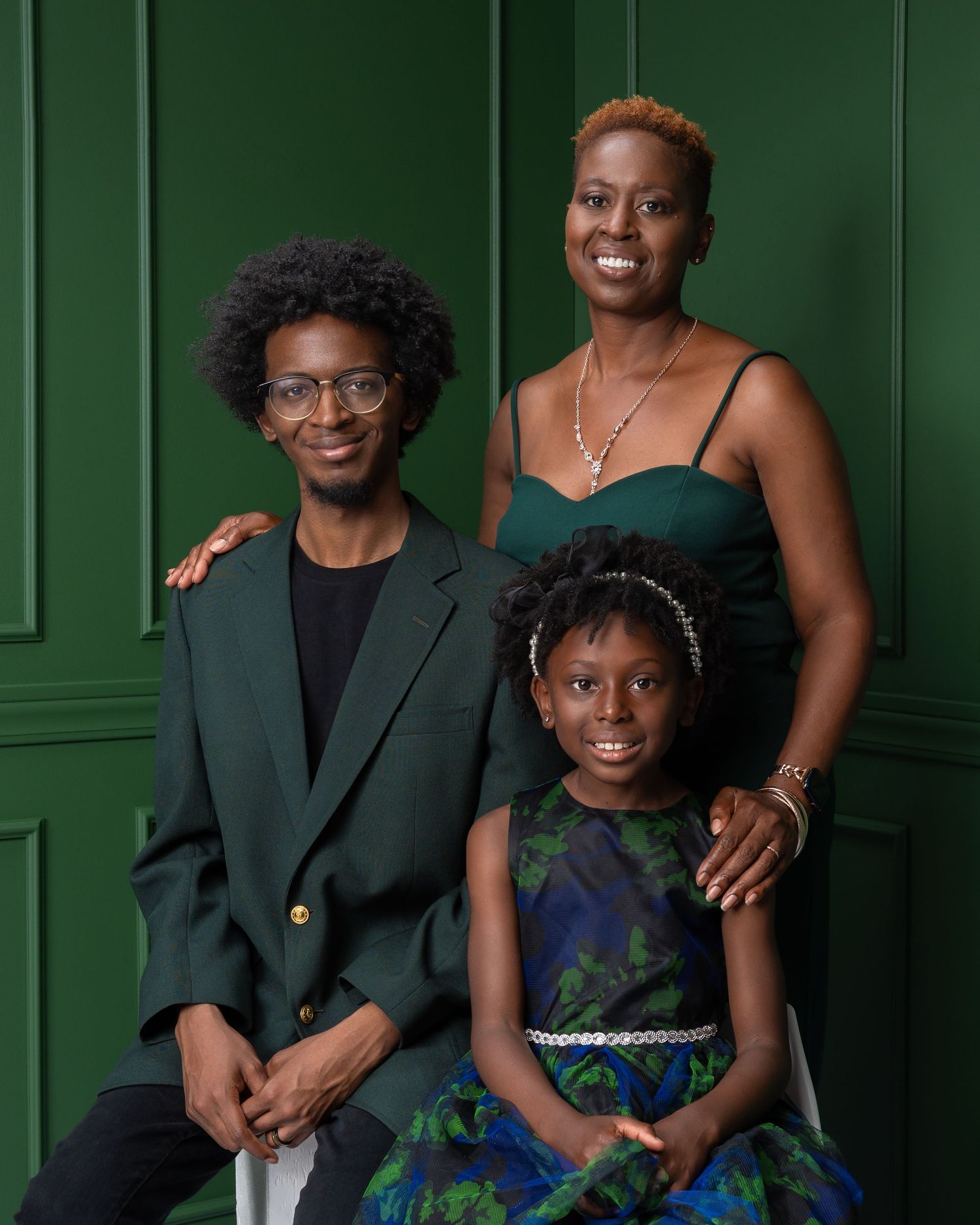 A joyful family of three poses in coordinating green outfits against an elegant green paneled wall.