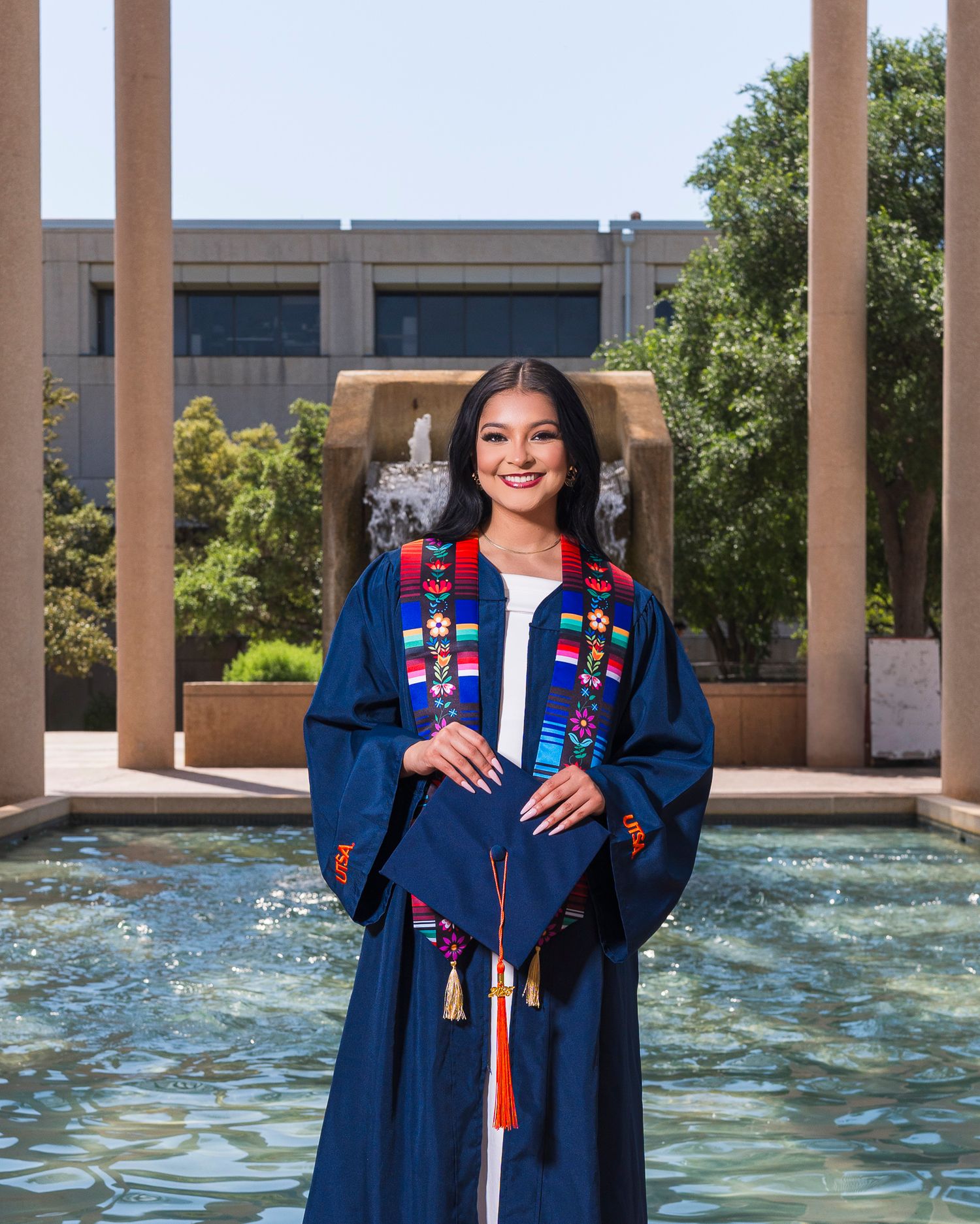 Graduate in navy blue gown with colorful embroidered stole holding cap near campus fountain.