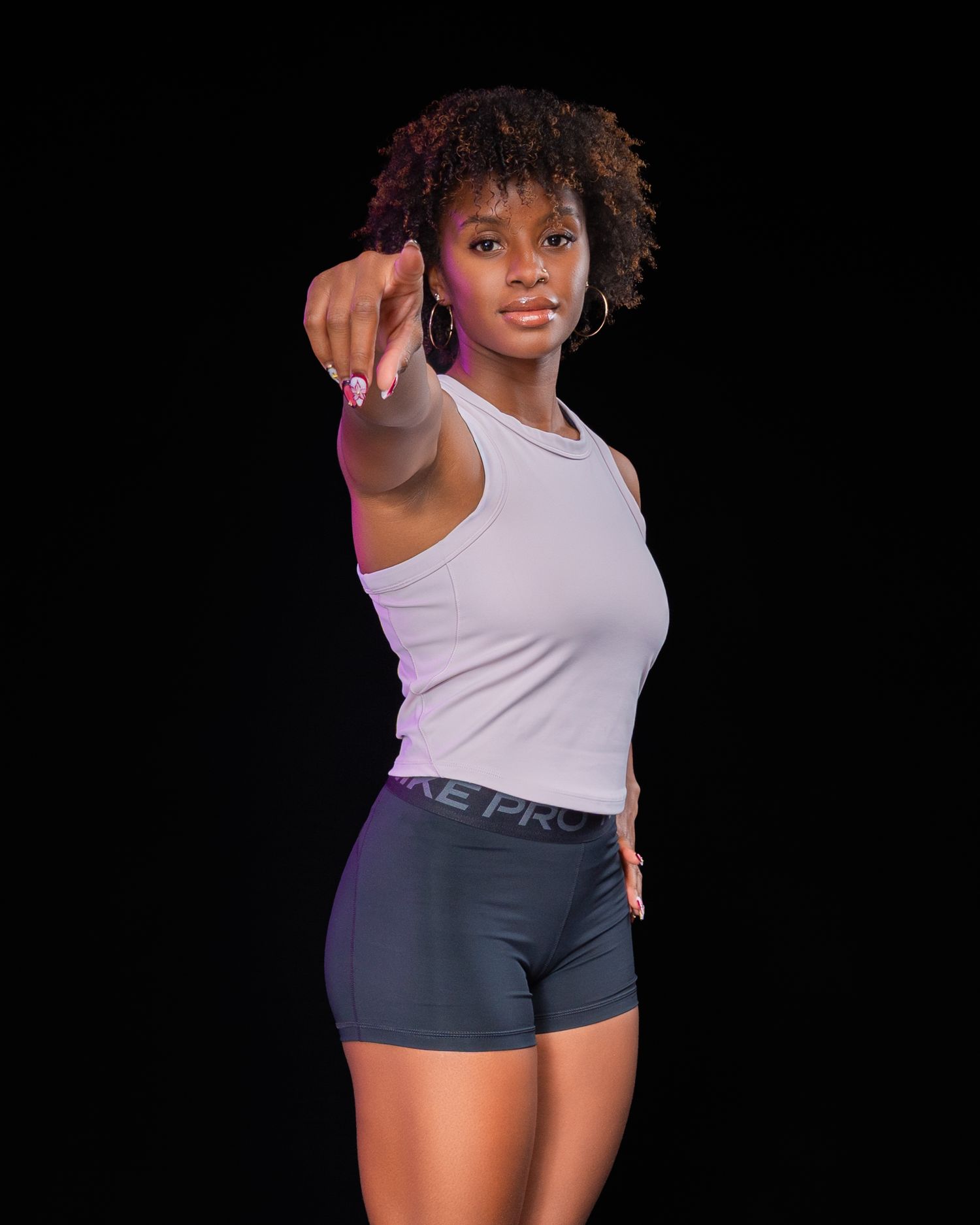 A fit Black woman in Nike Pro shorts and white tank top points directly at camera against black background.