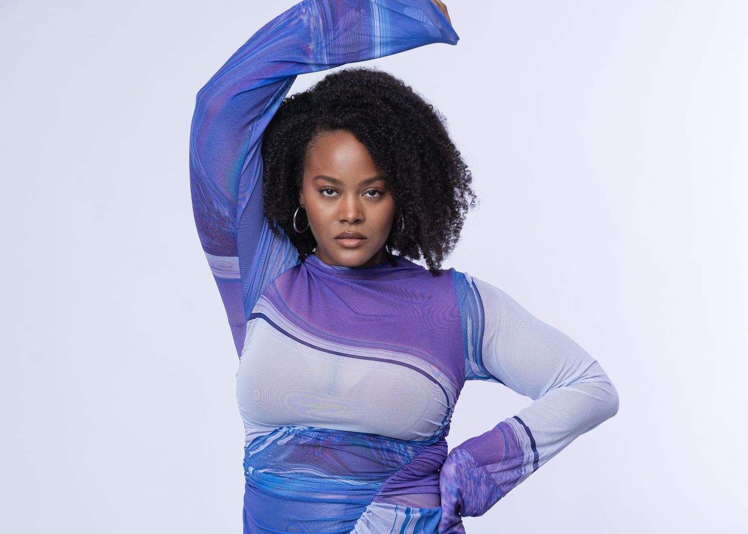 Woman in purple geometric mesh dress poses confidently against white background with flowing blue fabric.