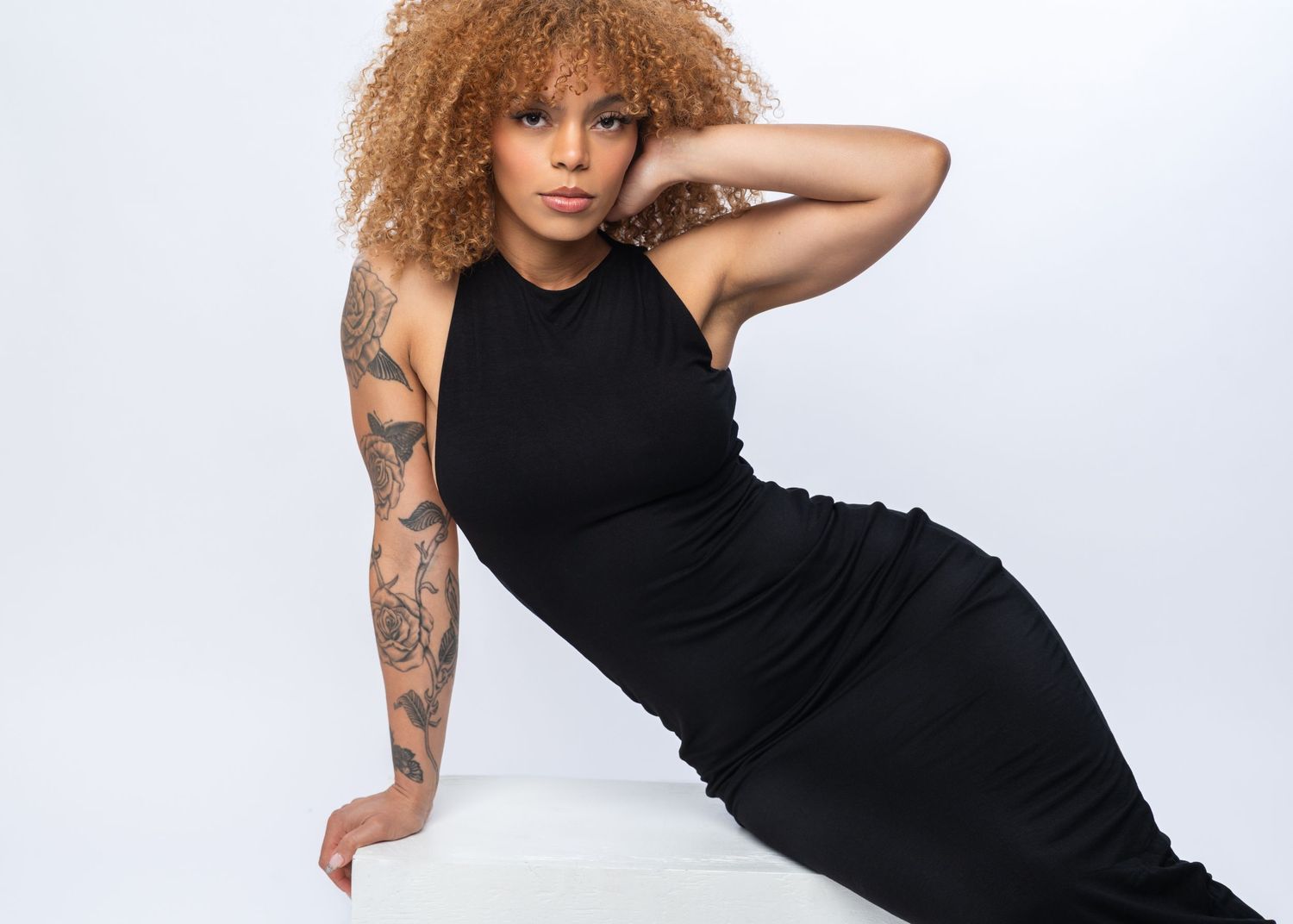 Woman in black dress with curly hair and tattoos posing confidently against a white background.