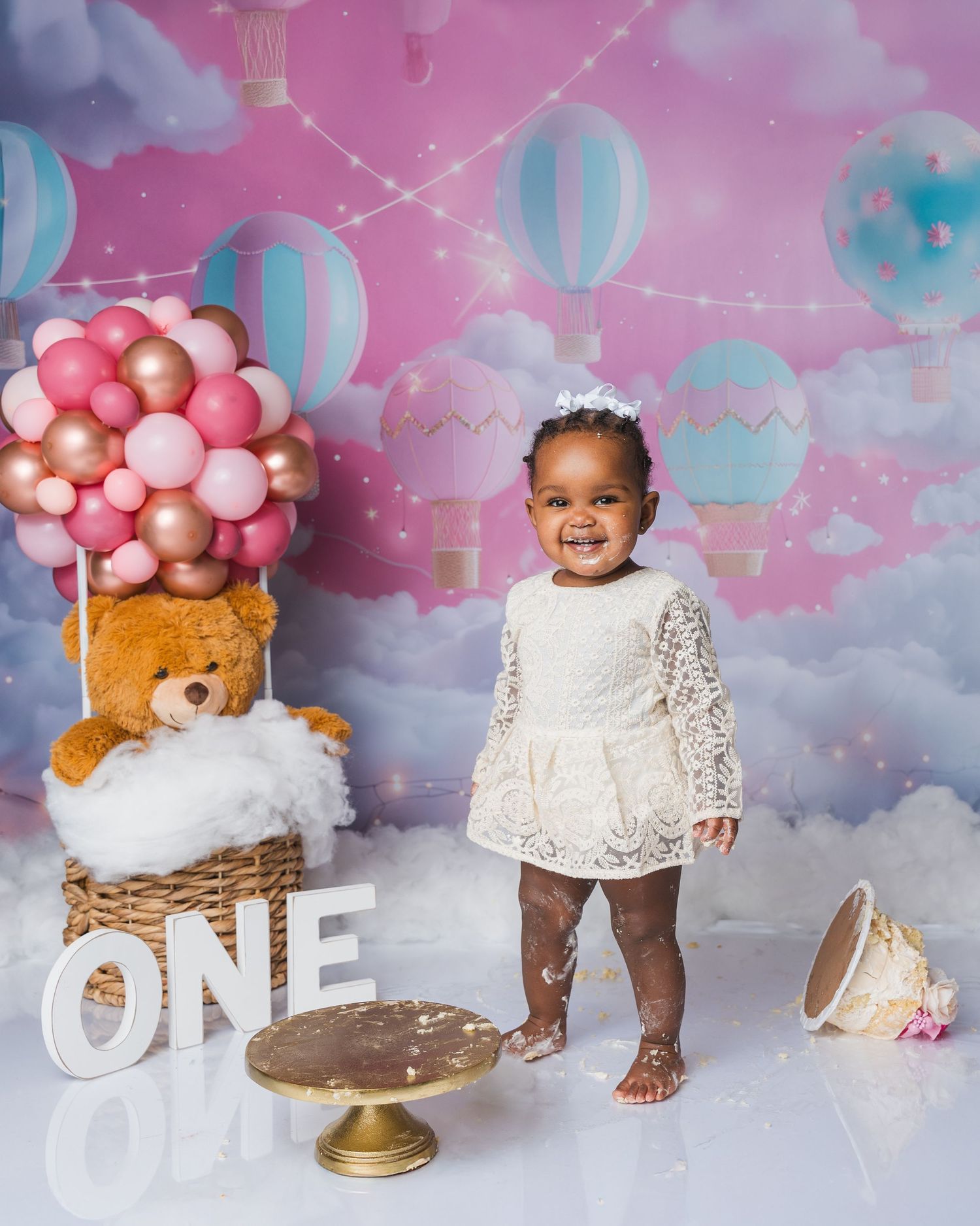 Smiling toddler in white lace dress celebrates first birthday with ONE sign, teddy bear balloon basket, and hot air balloon backdrop.