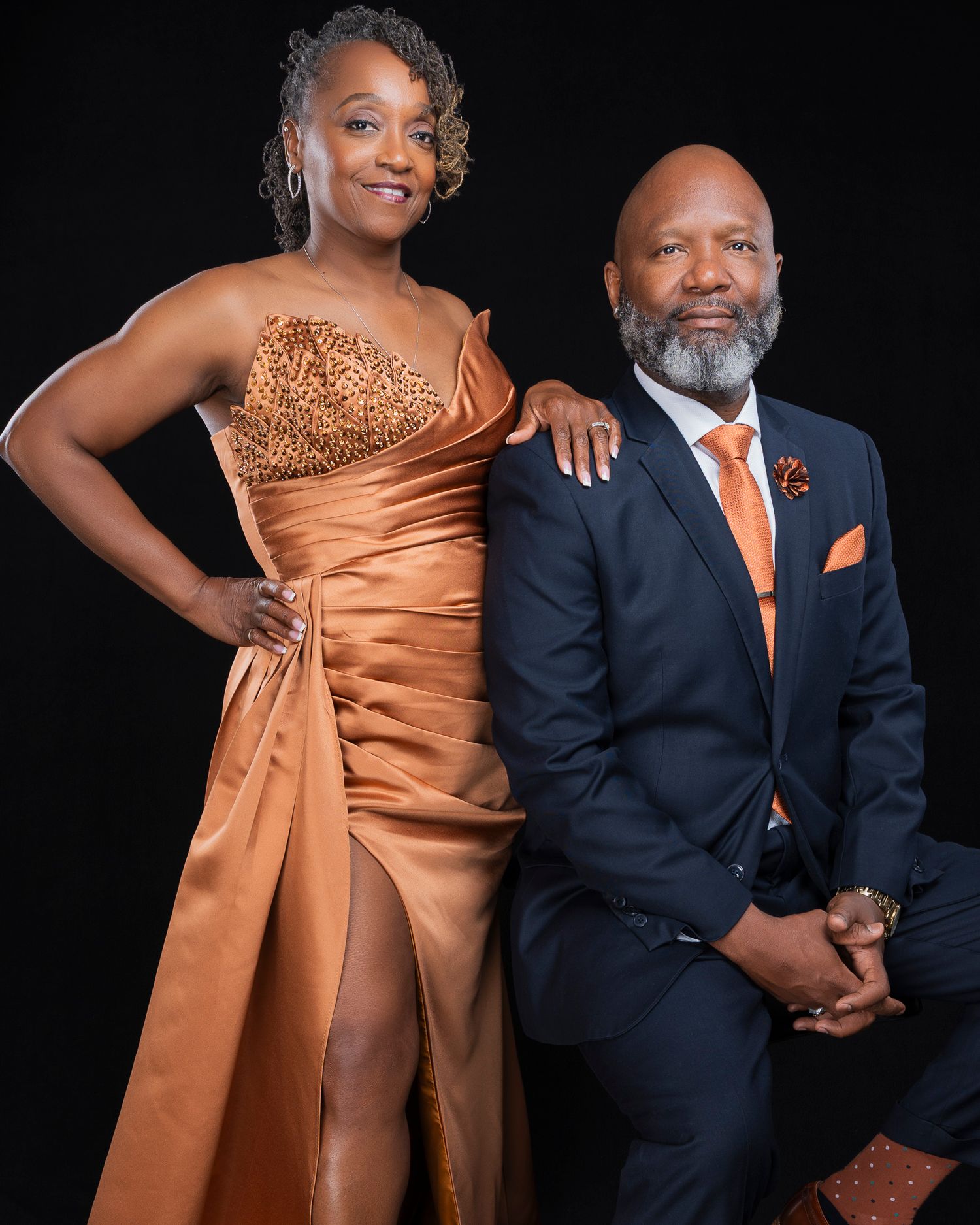 Sophisticated couple in matching orange gown and navy suit posing against dark studio background.