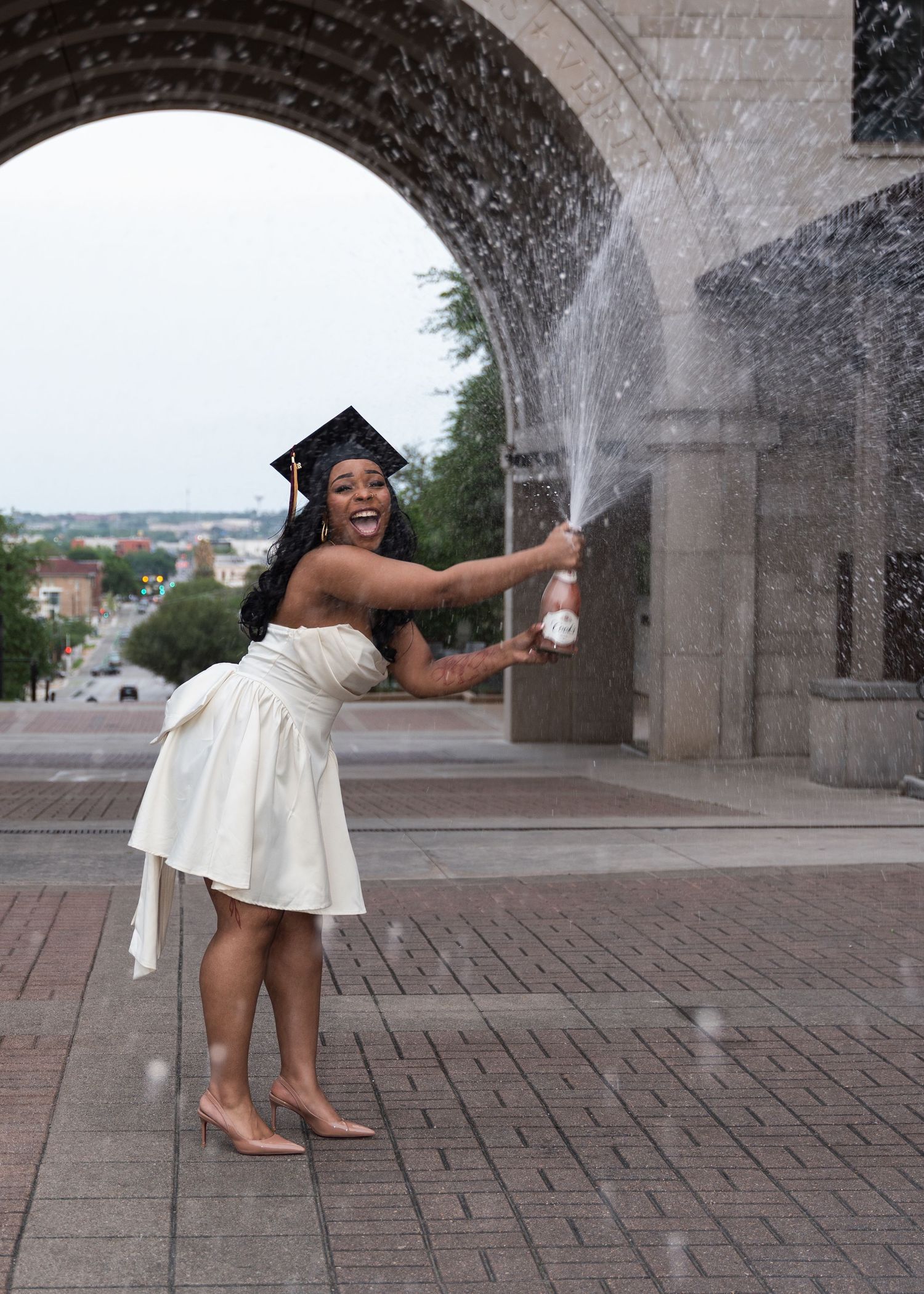 Graduate in white dress and cap sprays champagne bottle under stone arch, celebrating college graduation outdoors.