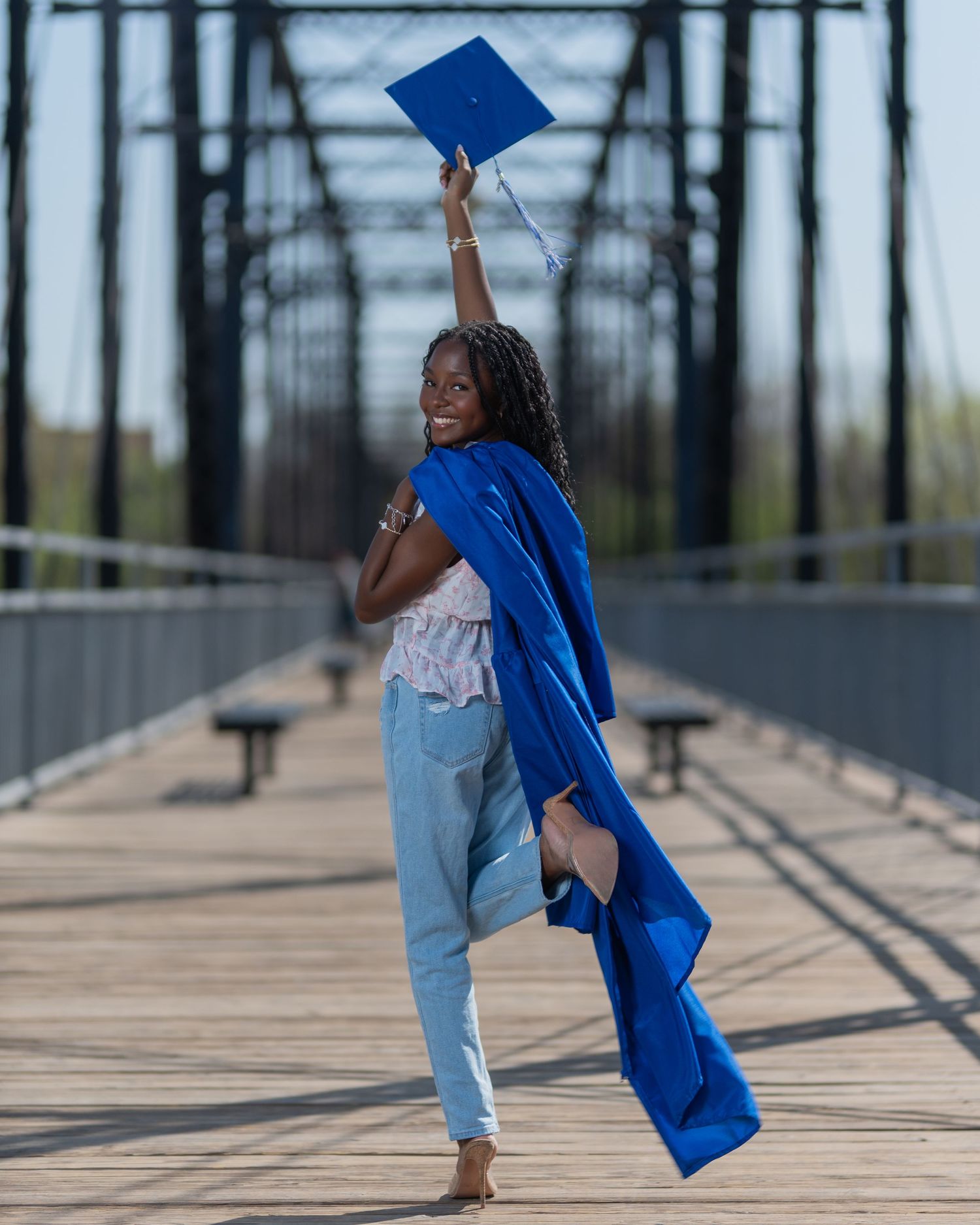 A joyful graduate in a blue cap and gown celebrates on a wooden bridge, holding her cap high in the air.