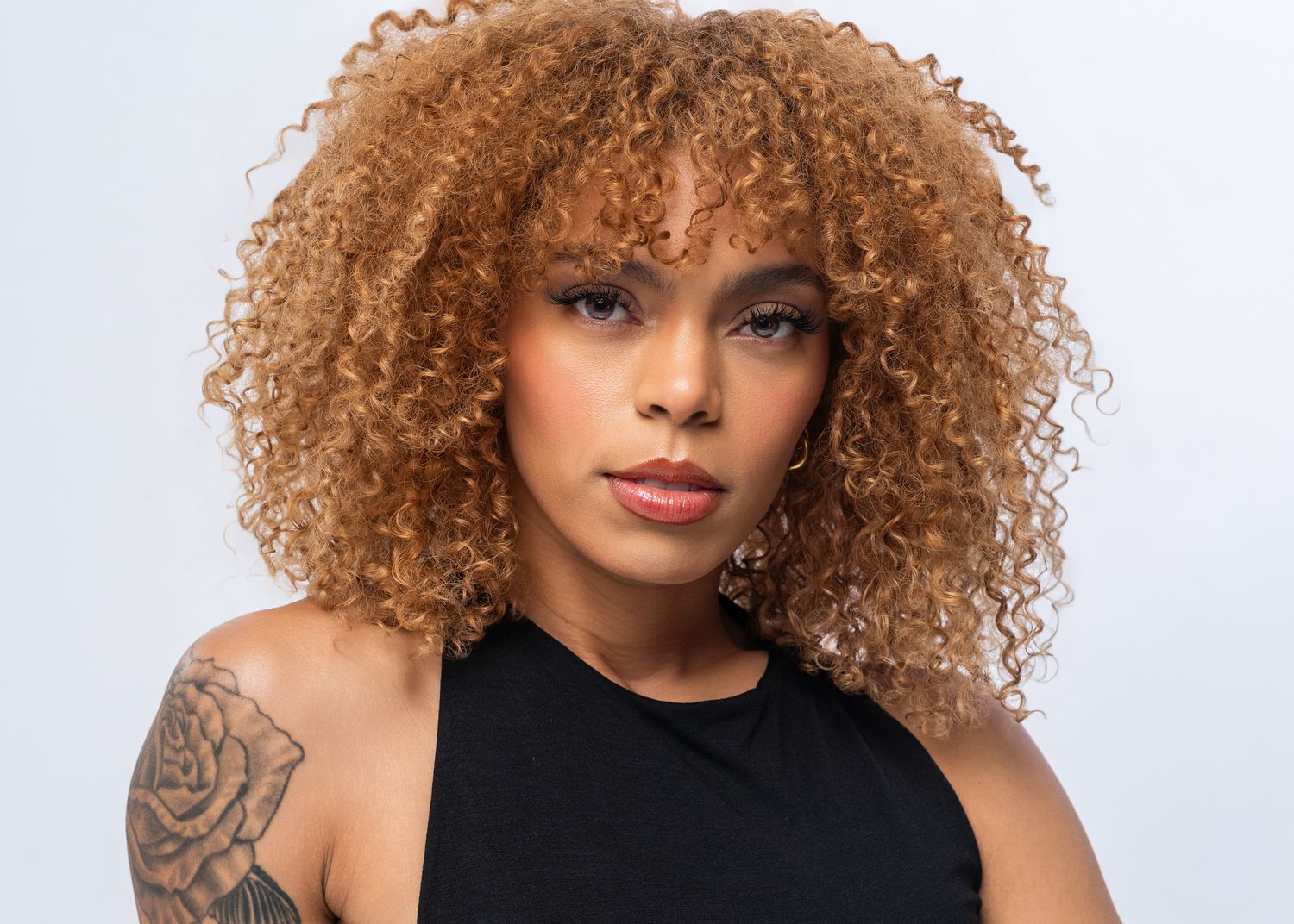 Young woman with curly auburn hair, tattoos on shoulder, wearing black top, posing against white background.