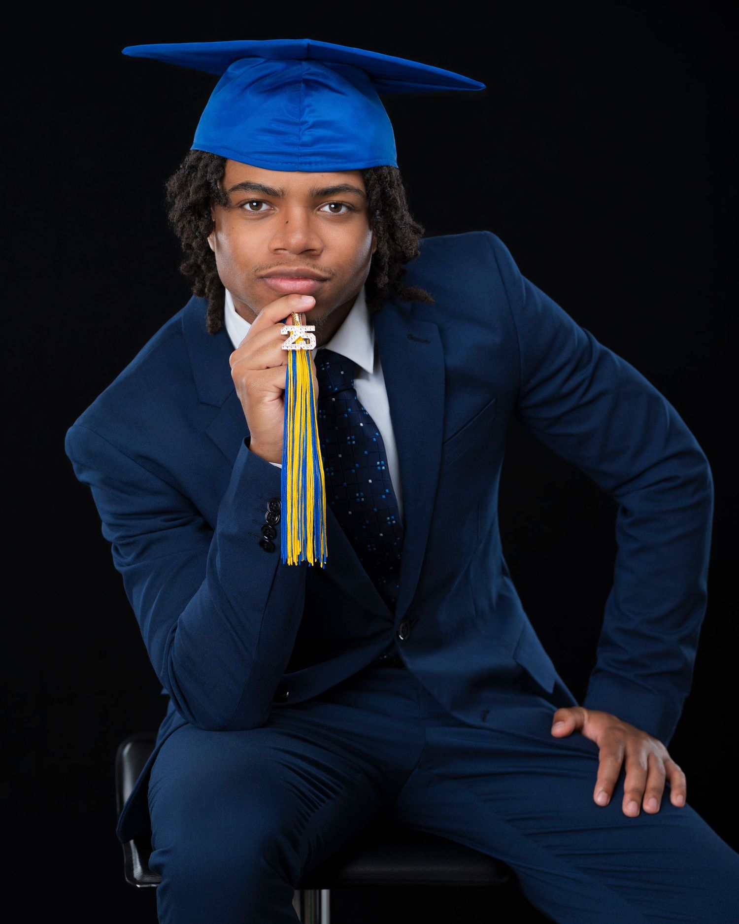 Young man in blue suit and graduation cap holds tassel, posing confidently for senior portrait on dark background.