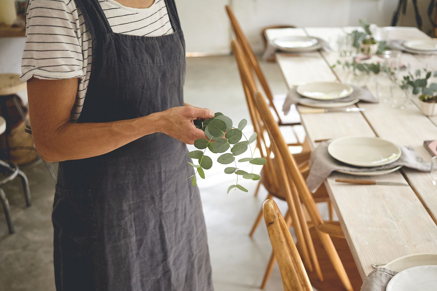Small business owner in a linen apron holding a eucalyptus sprig beside a styled table during a brand photoshoot