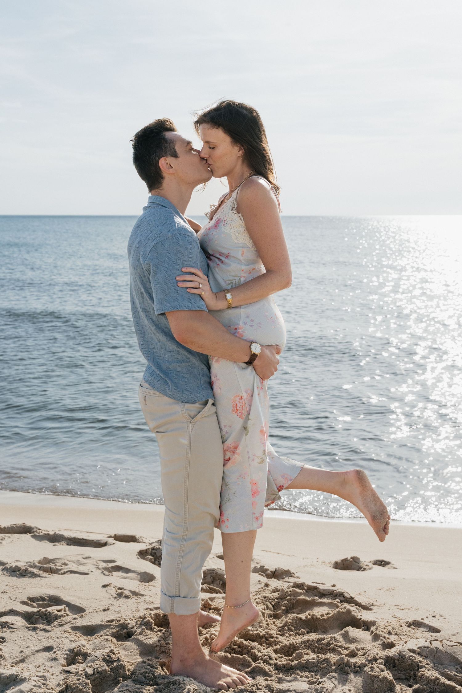 Couple sharing a kiss on a sunny beach, woman in floral dress with one leg raised, lake backdrop.