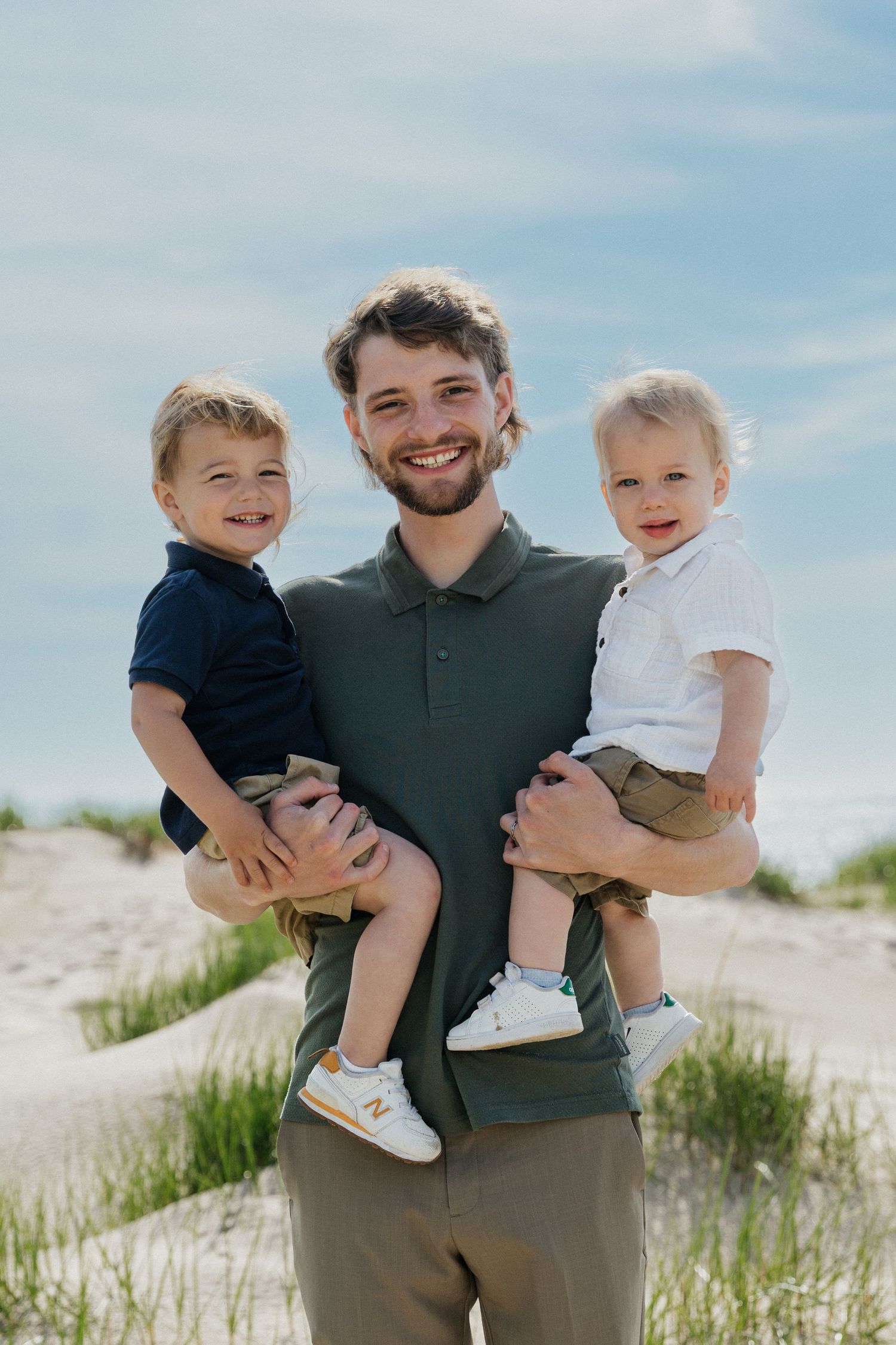 Smiling dad holding two toddlers on a sandy beach with grass dunes and blue sky backdrop.