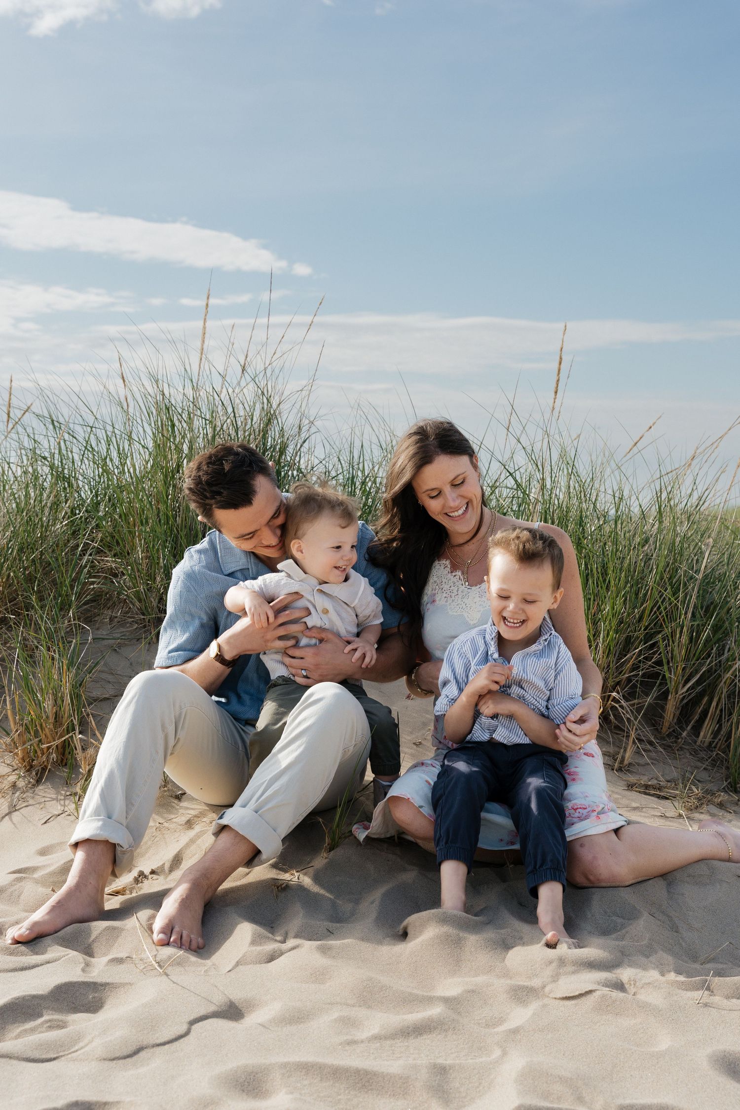 Family of four sitting on sandy beach dunes, parents smiling at baby and young boy, with tall grass and blue sky behind.