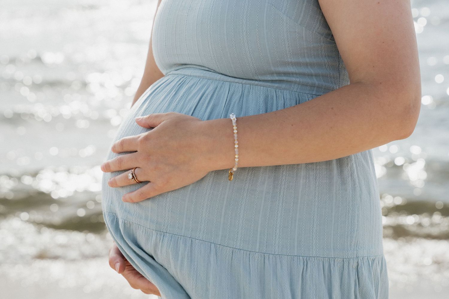 Pregnant woman in a light blue dress cradling her baby bump near a shimmering ocean, wearing a delicate gold bracelet.