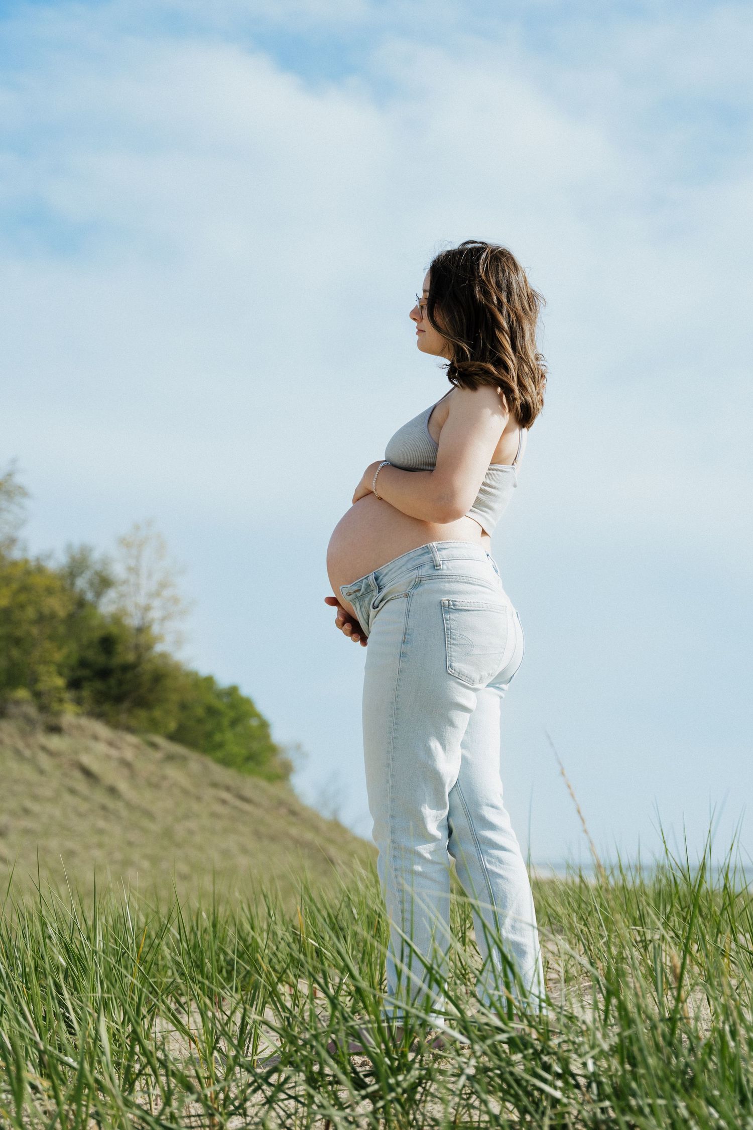 Pregnant woman in jeans and crop top standing outdoors on sandy dune with green grass and blue sky.
