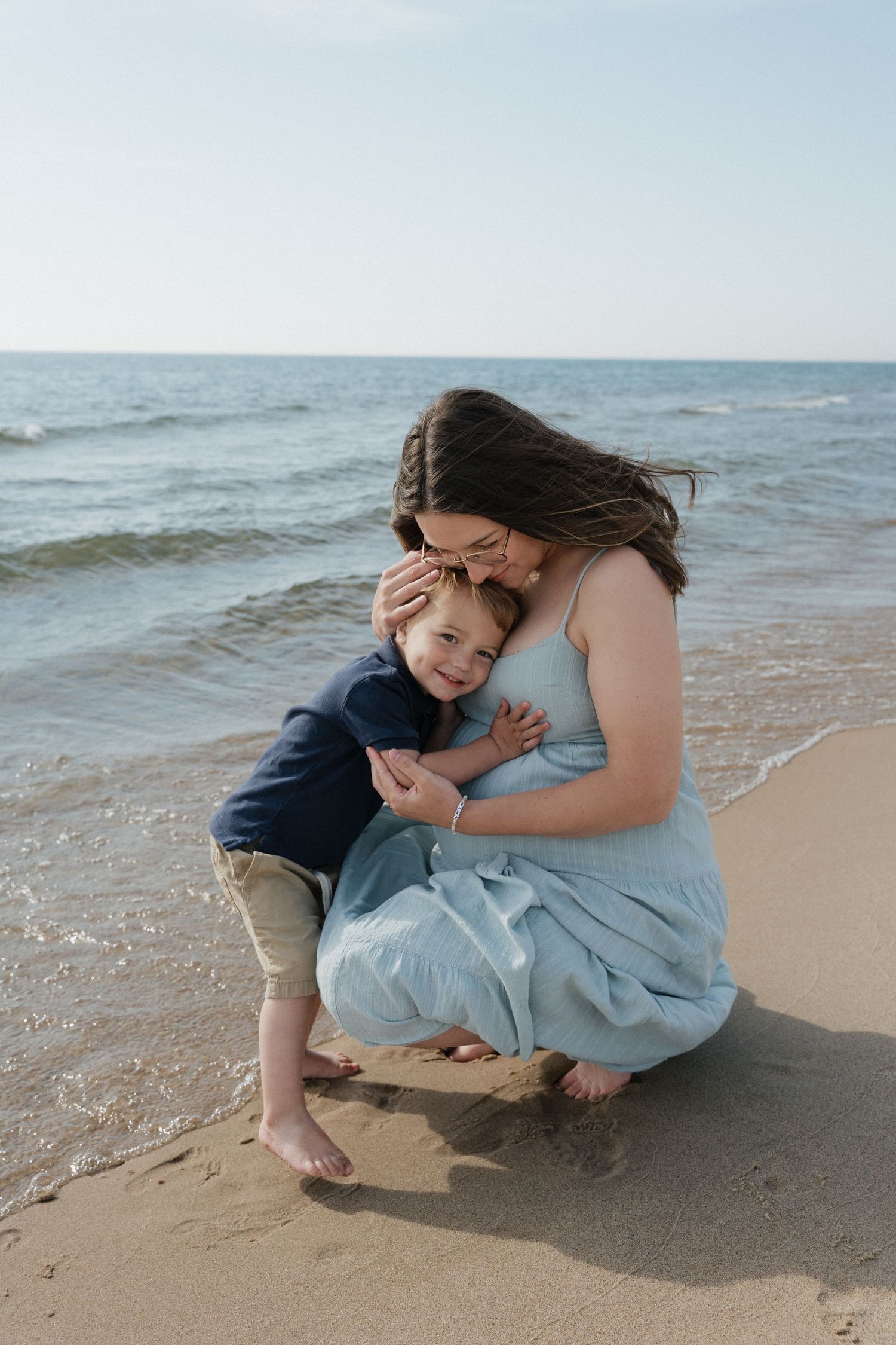 A mother in a light blue dress crouches on a sandy beach, embracing her young son near the ocean waves.