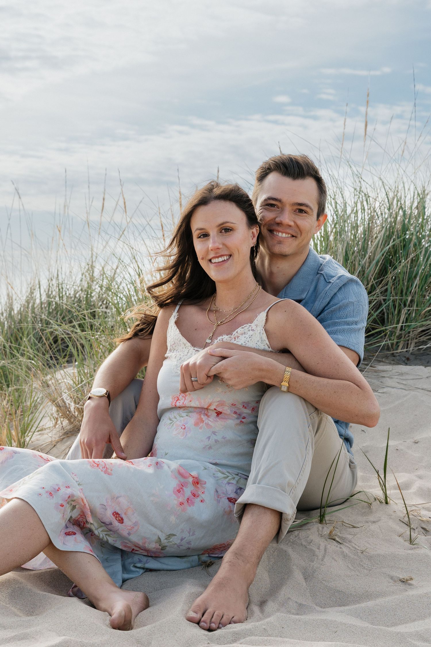 A smiling couple sits together on sandy beach dunes, the woman in a floral dress leaning back against the man in a blue shirt.