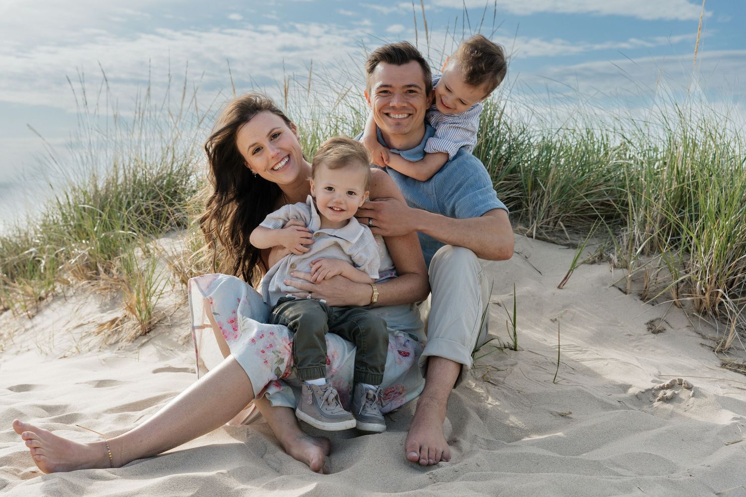 Happy family of four smiling together on sandy beach dunes with ocean backdrop on a sunny day.