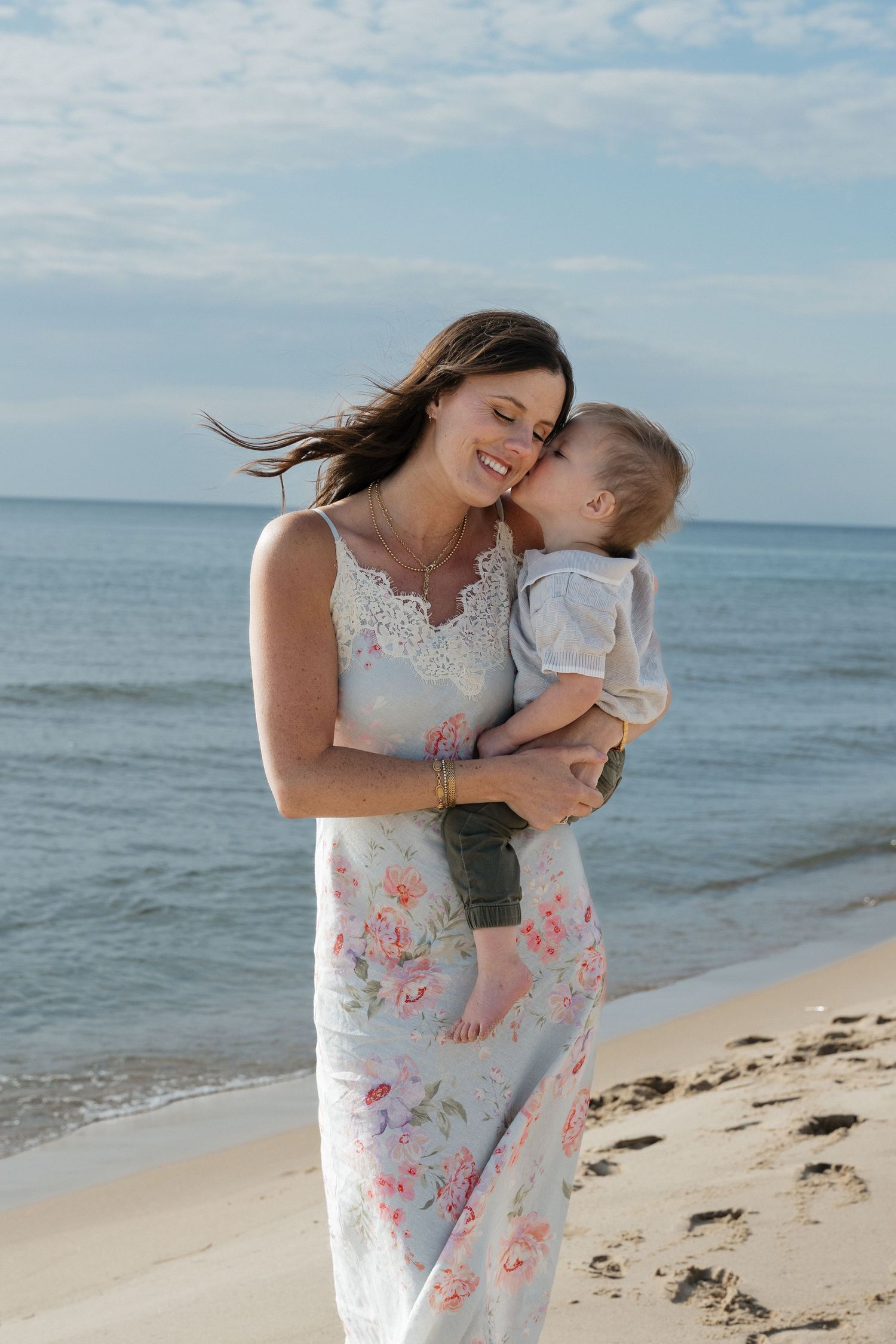 A smiling woman in a floral dress holds a baby on a sandy beach with waves in the background.