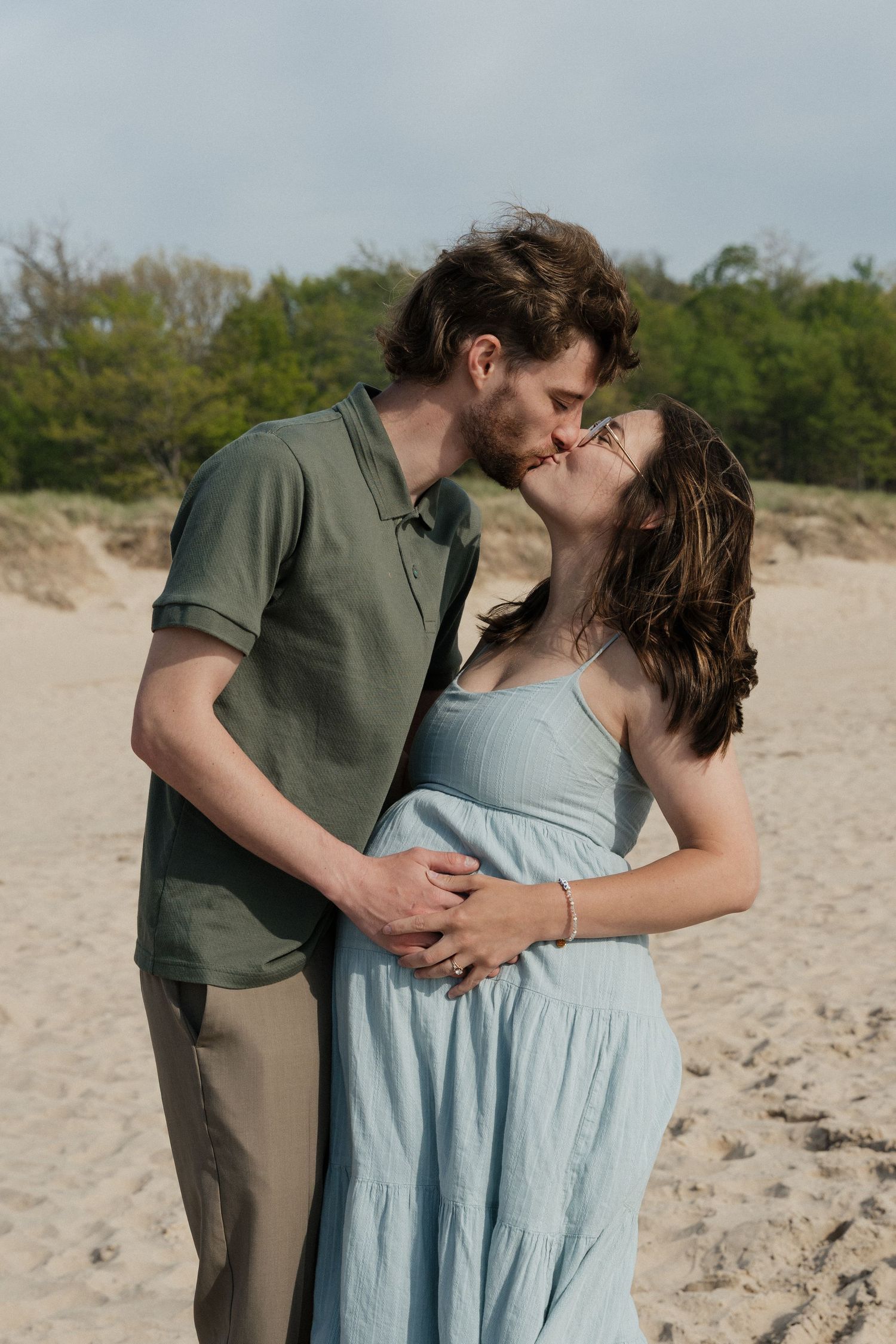 A couple shares a romantic kiss on a sandy beach, the woman in a blue sundress and man in a green shirt embracing tenderly.