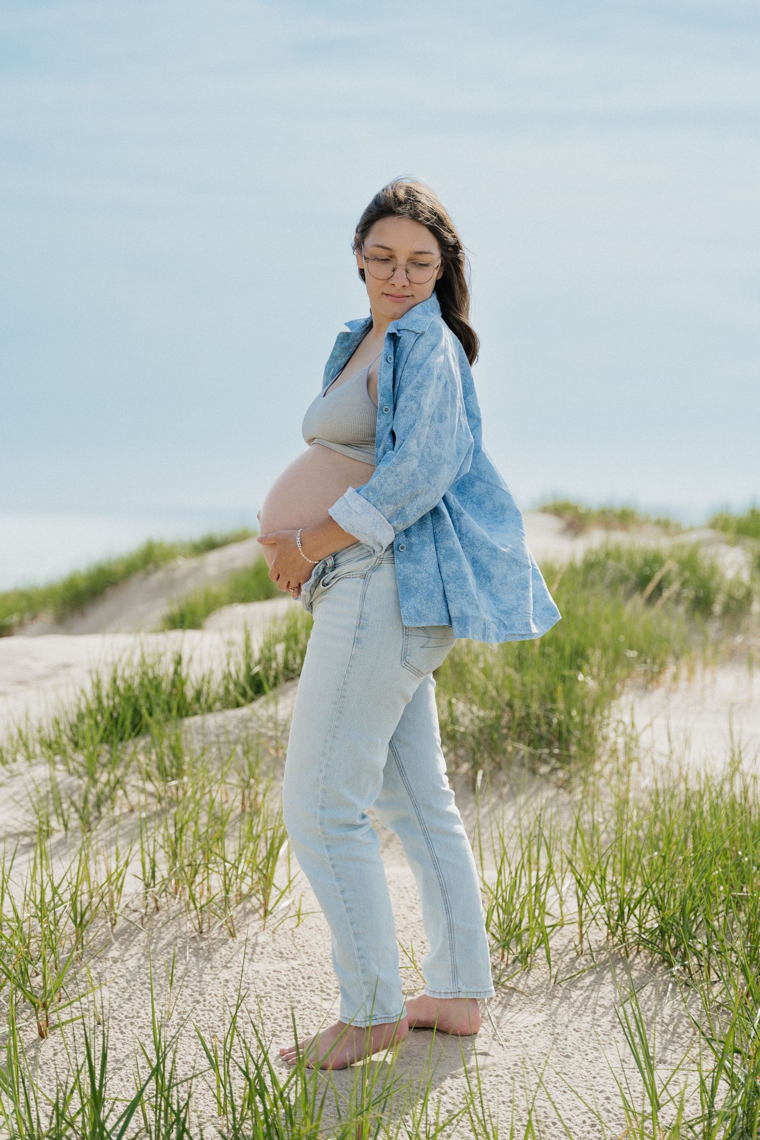 Pregnant woman in jeans and blue shirt standing barefoot on sandy beach dunes, cradling her baby bump outdoors.