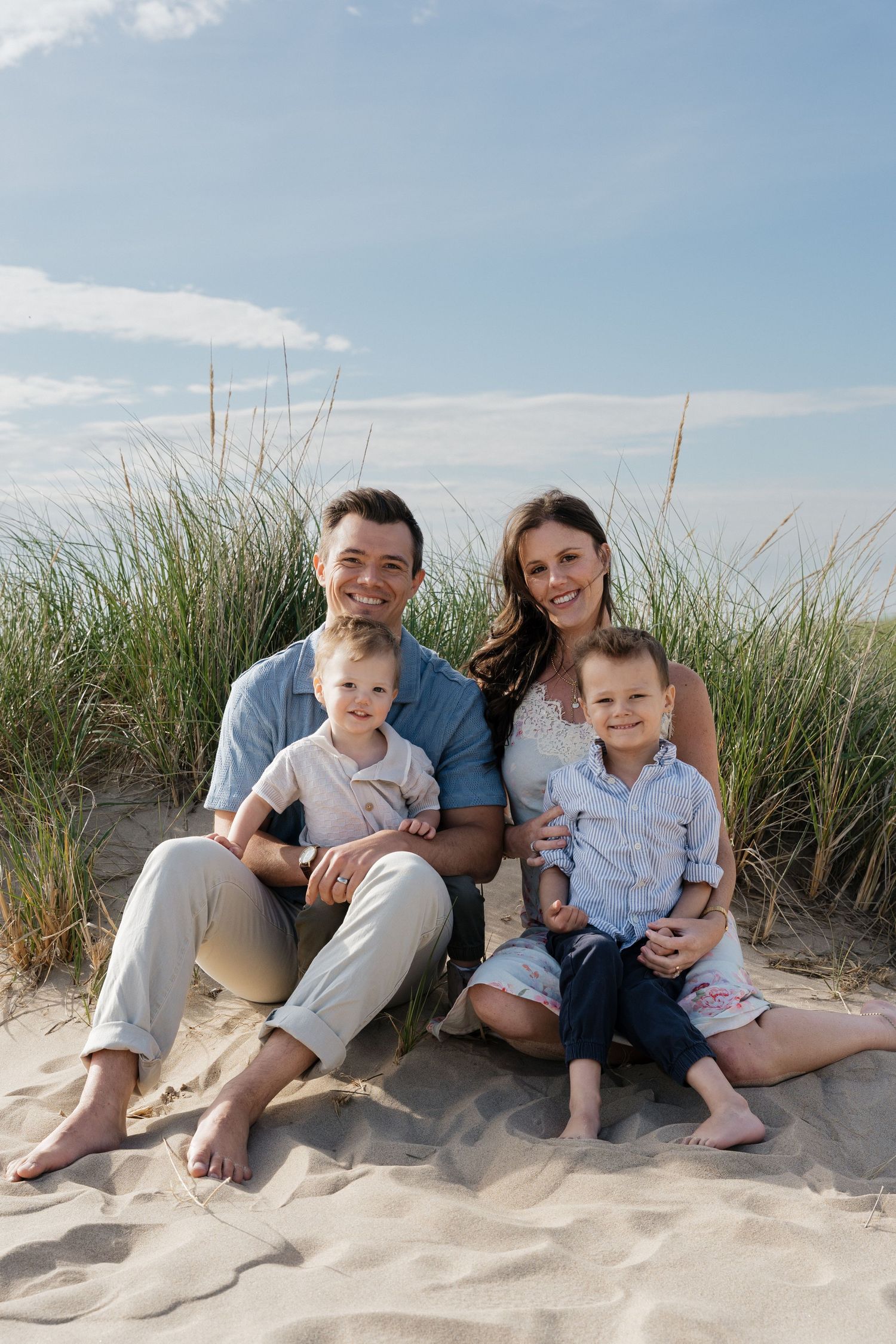 Family of four smiling together on sandy beach dunes with green grass and blue sky backdrop.