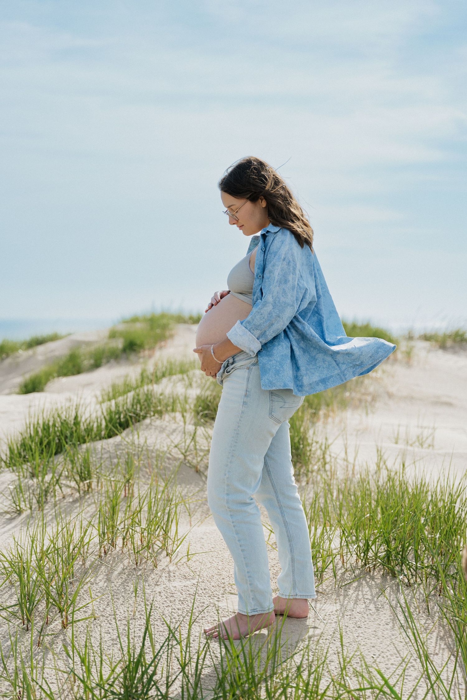 Pregnant woman in blue shirt and jeans cradling her baby bump on sandy beach dunes with green grass.