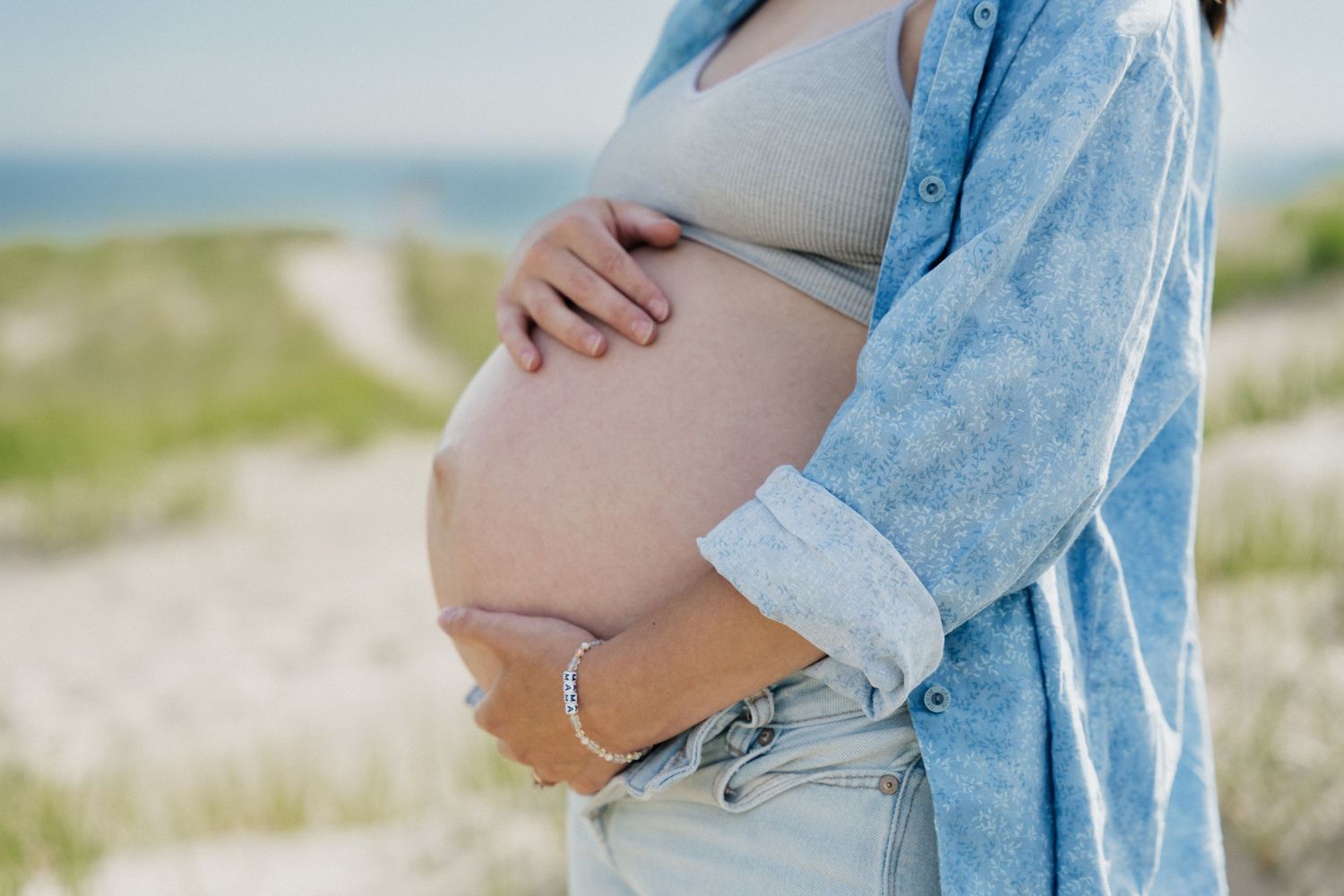 Pregnant woman in blue shirt and jeans cradling baby bump at sandy beach on a sunny day.
