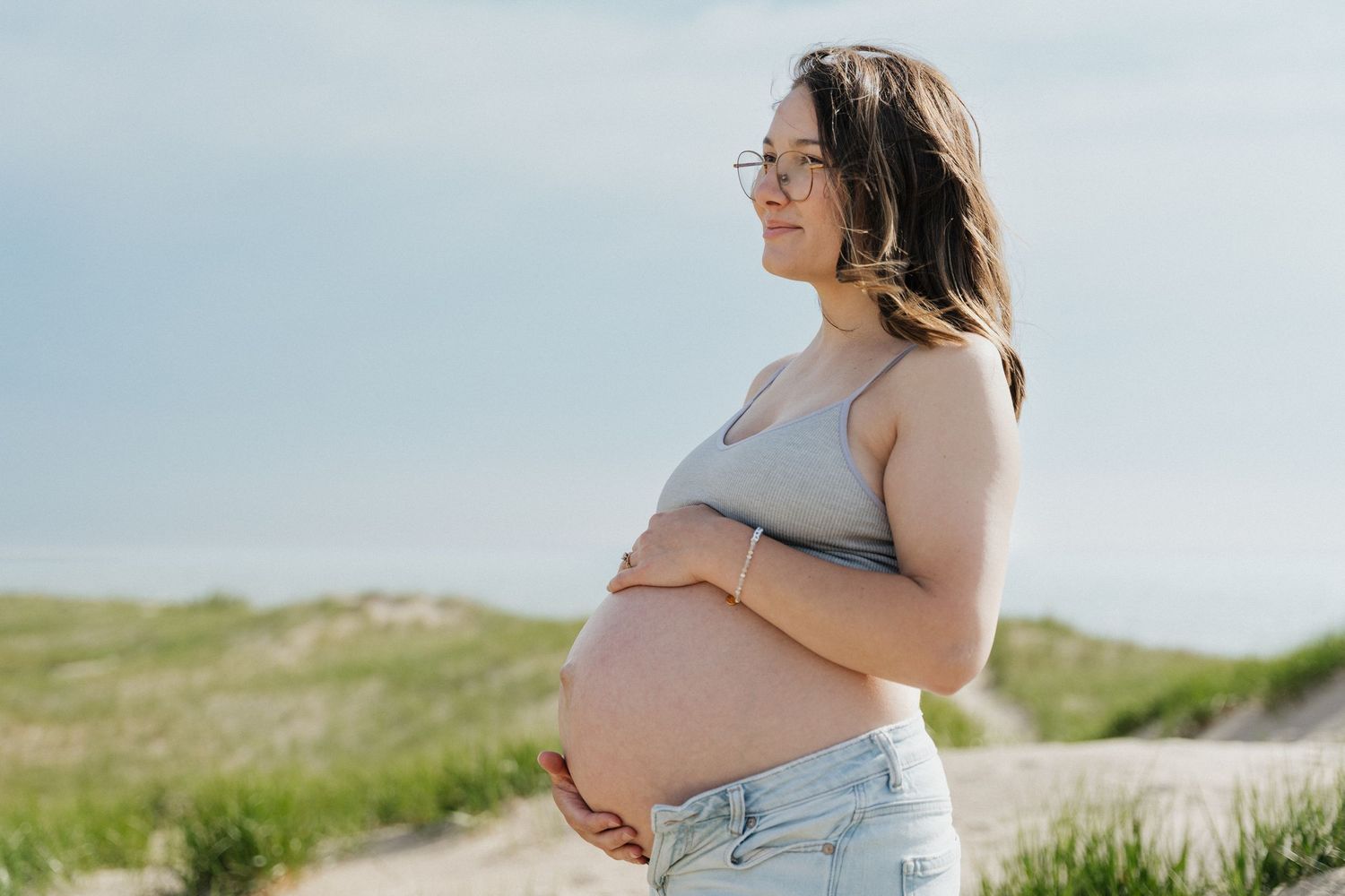 Pregnant woman in gray crop top and jeans holding baby bump on sandy beach, smiling profile view outdoors.