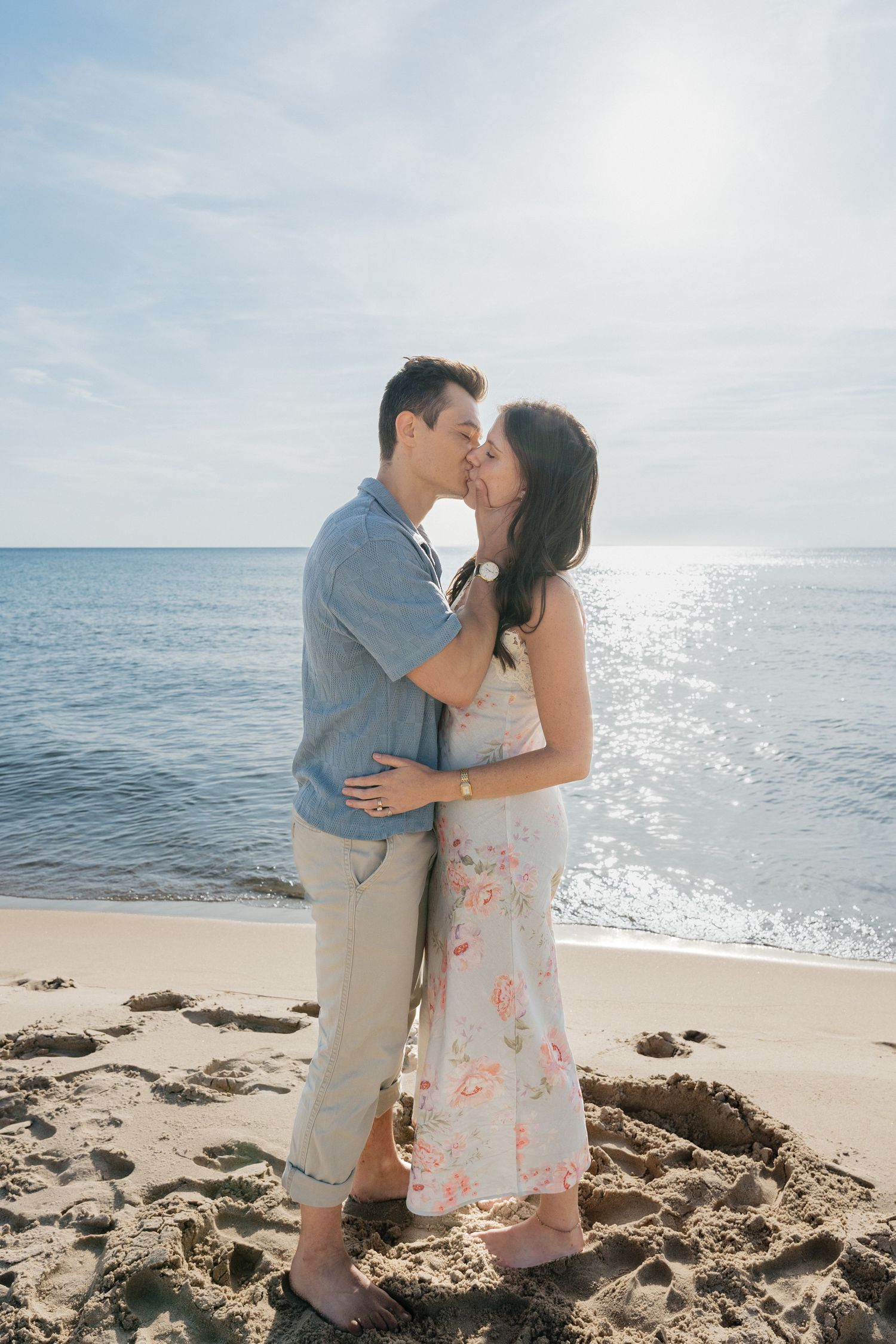 A couple shares a romantic kiss on a sunny beach, with sparkling waves and blue sky in the background.