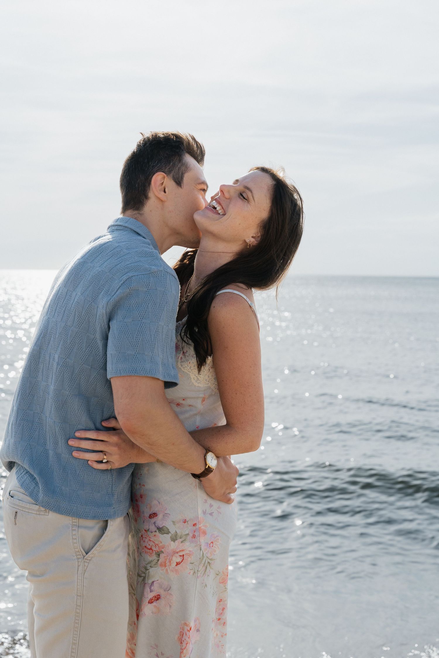 Romantic couple embracing and kissing on a beach with sparkling water in the background.