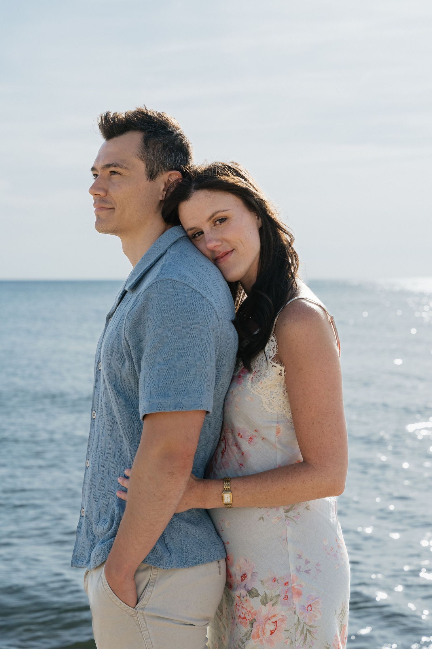 Couple embracing by the lake, woman in floral dress leaning on man in blue shirt, romantic seaside portrait.