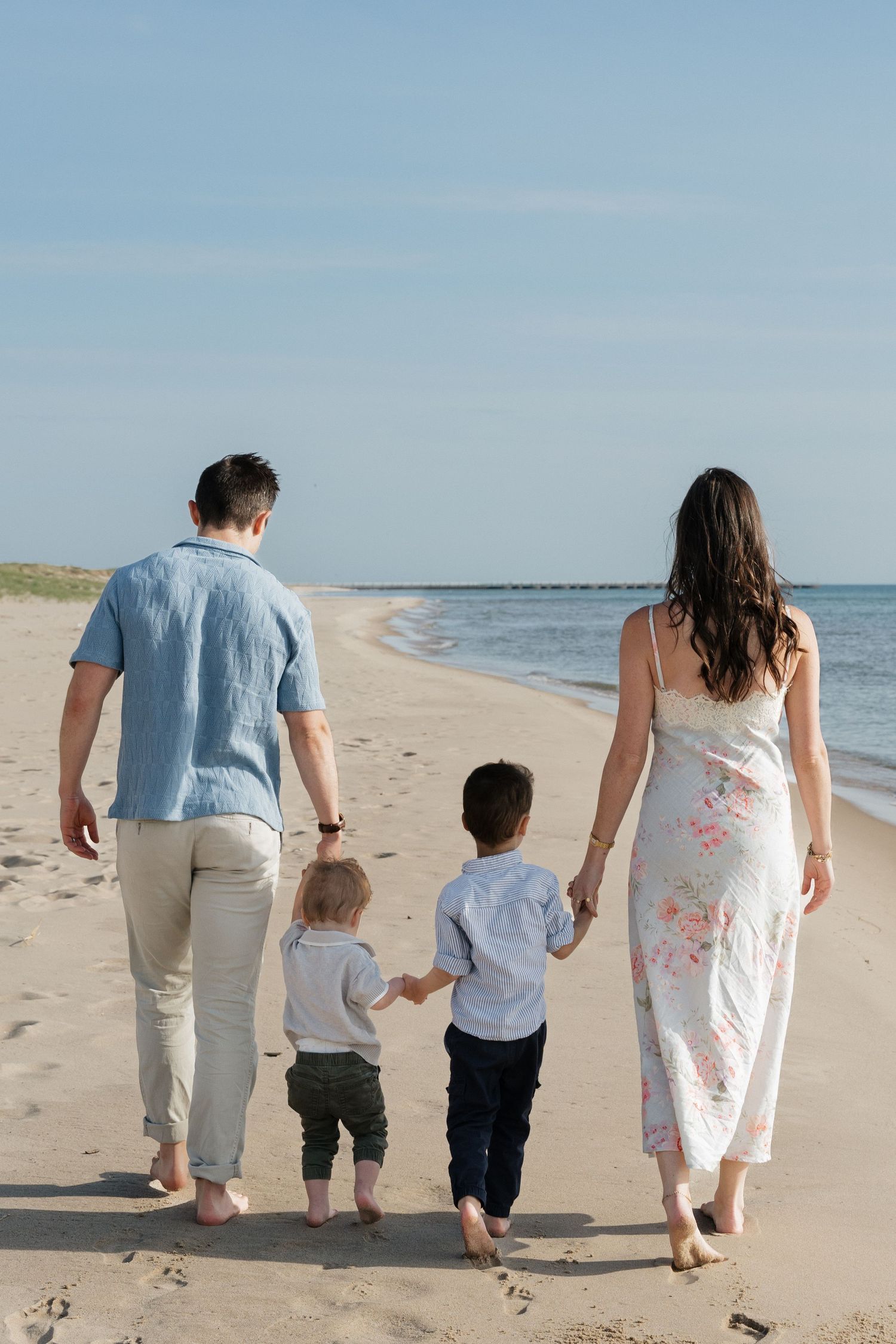 Family of four walking hand-in-hand on a sandy beach toward the ocean on a sunny day.