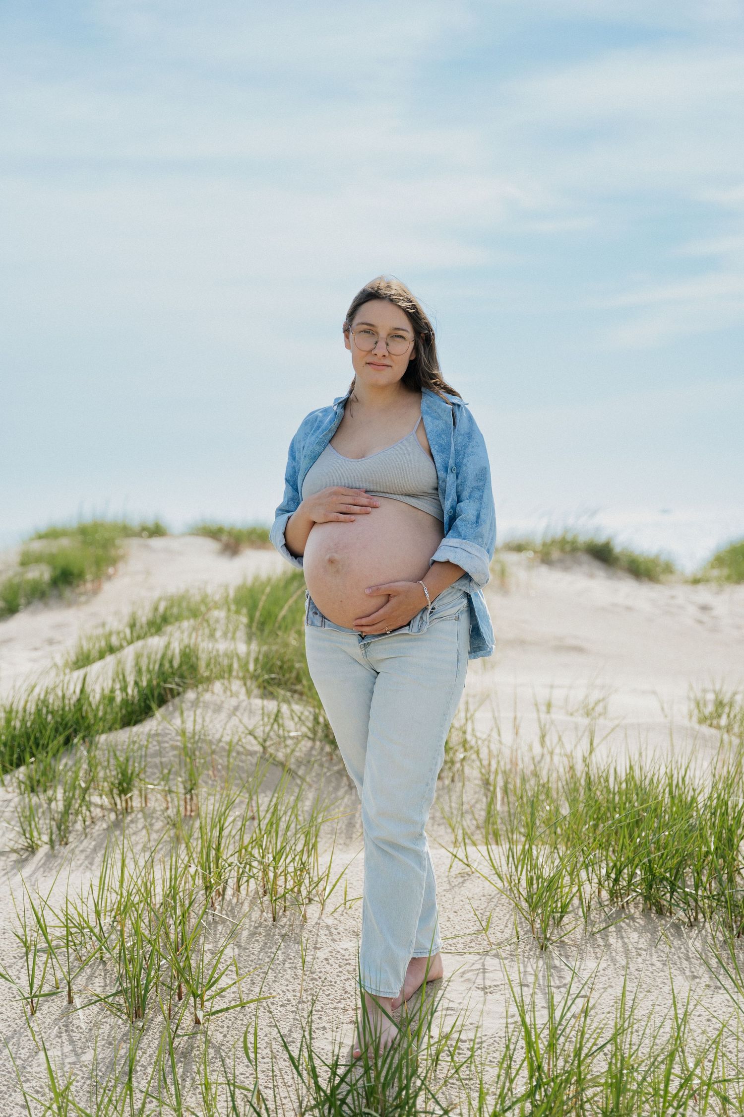 Pregnant woman in denim shirt and jeans cradling her baby bump on sandy beach dunes with green grass.