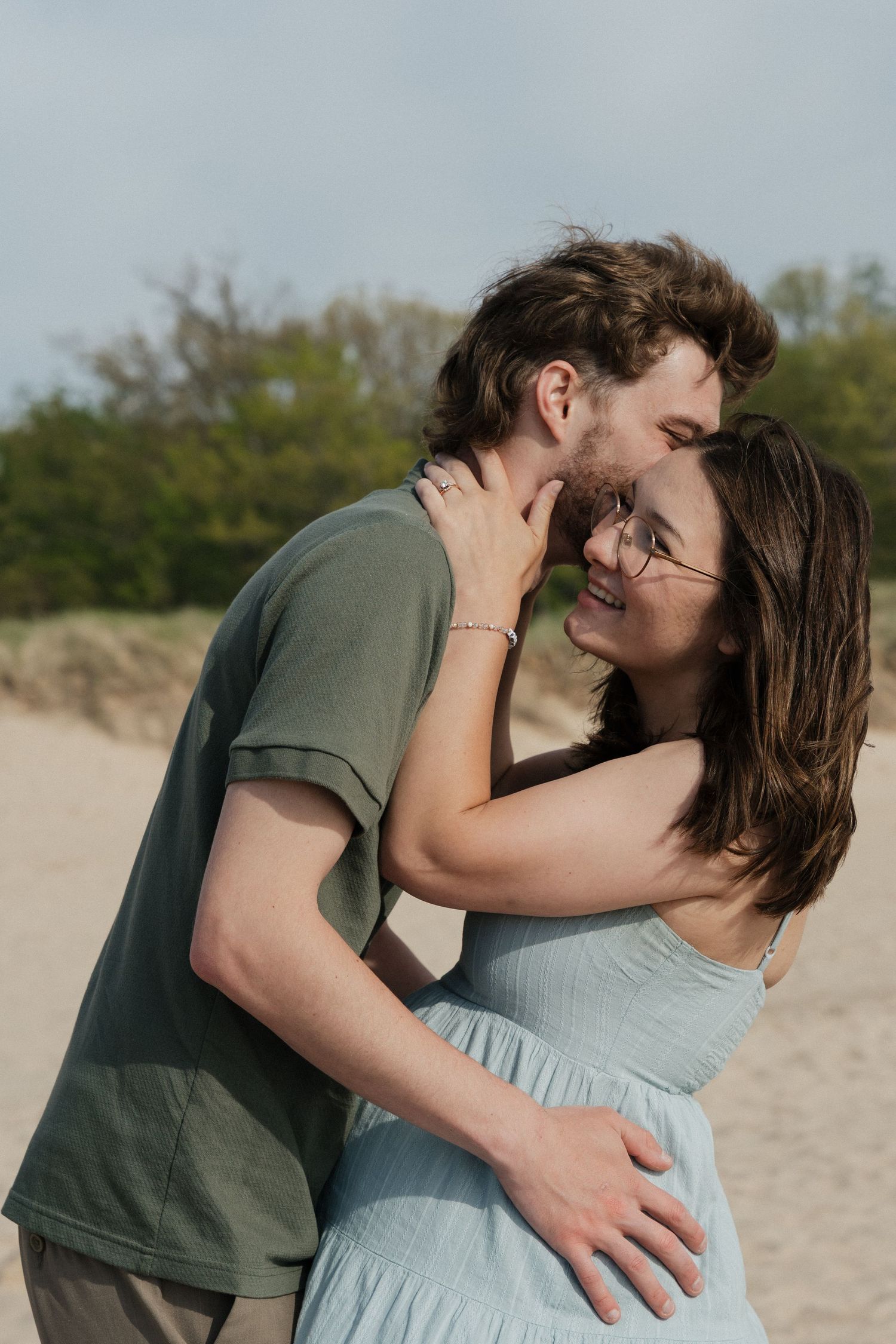 A couple embracing and smiling on a sandy beach, the woman in a blue dress and man in a green shirt, with trees behind.