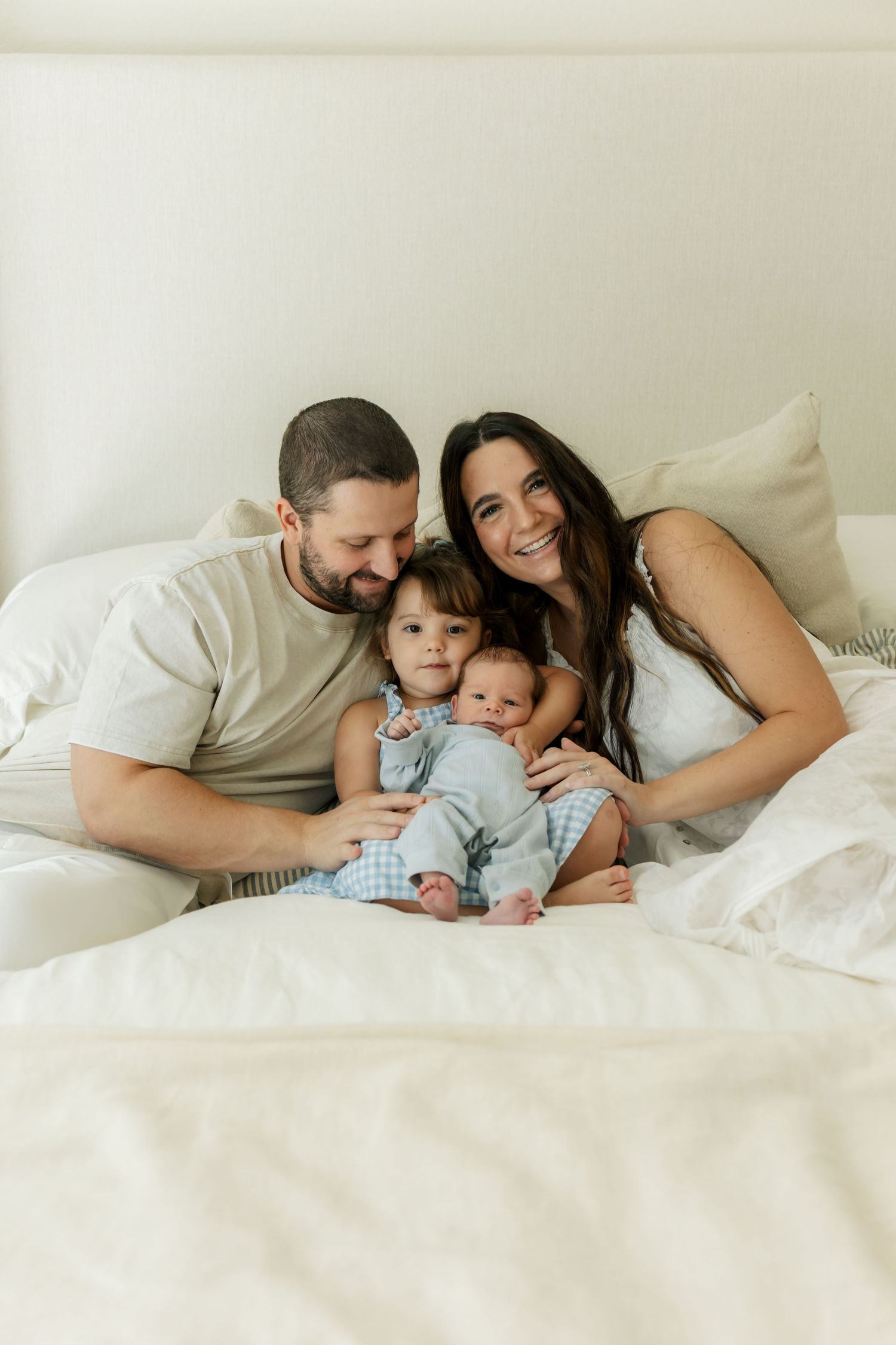 Happy family of four with parents and two young children snuggling together on a white bed in a bright room.