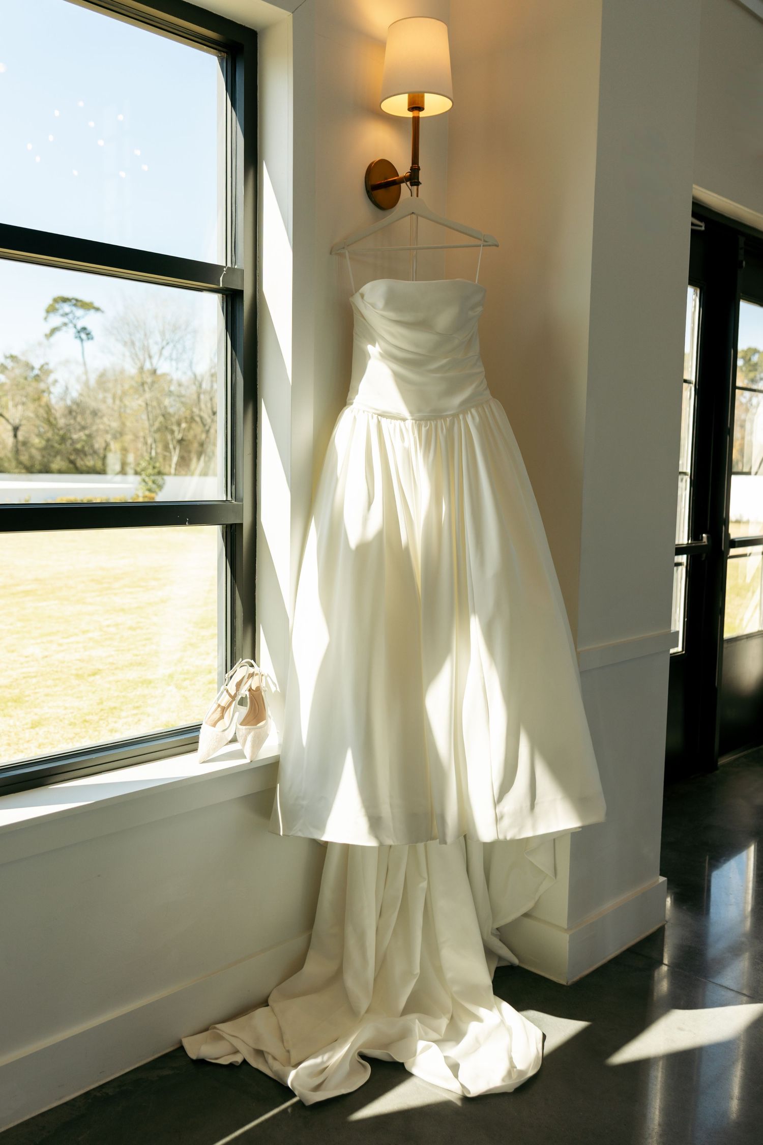 Elegant strapless white wedding gown with long train hanging on wall hanger near sunlit window indoors.