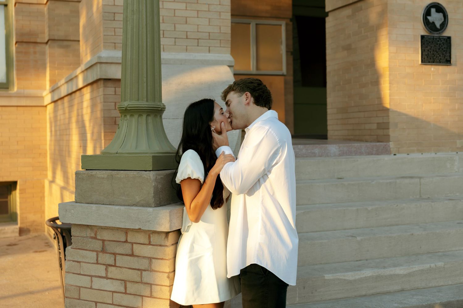 Couple sharing a kiss beside a column of a sunlit historic brick building.