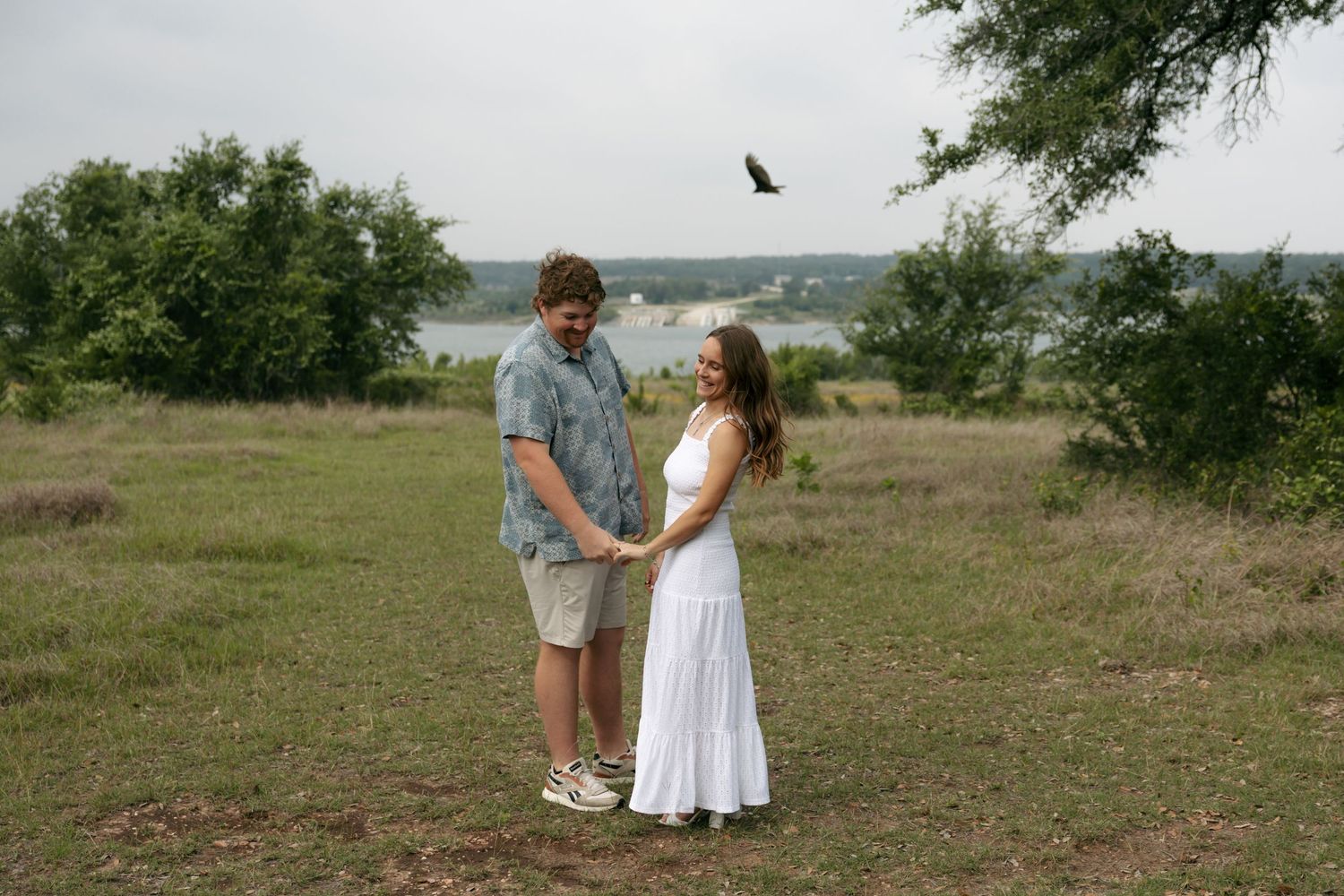 Couple holds hands in a scenic lakeside field with a bird soaring in the cloudy sky above.
