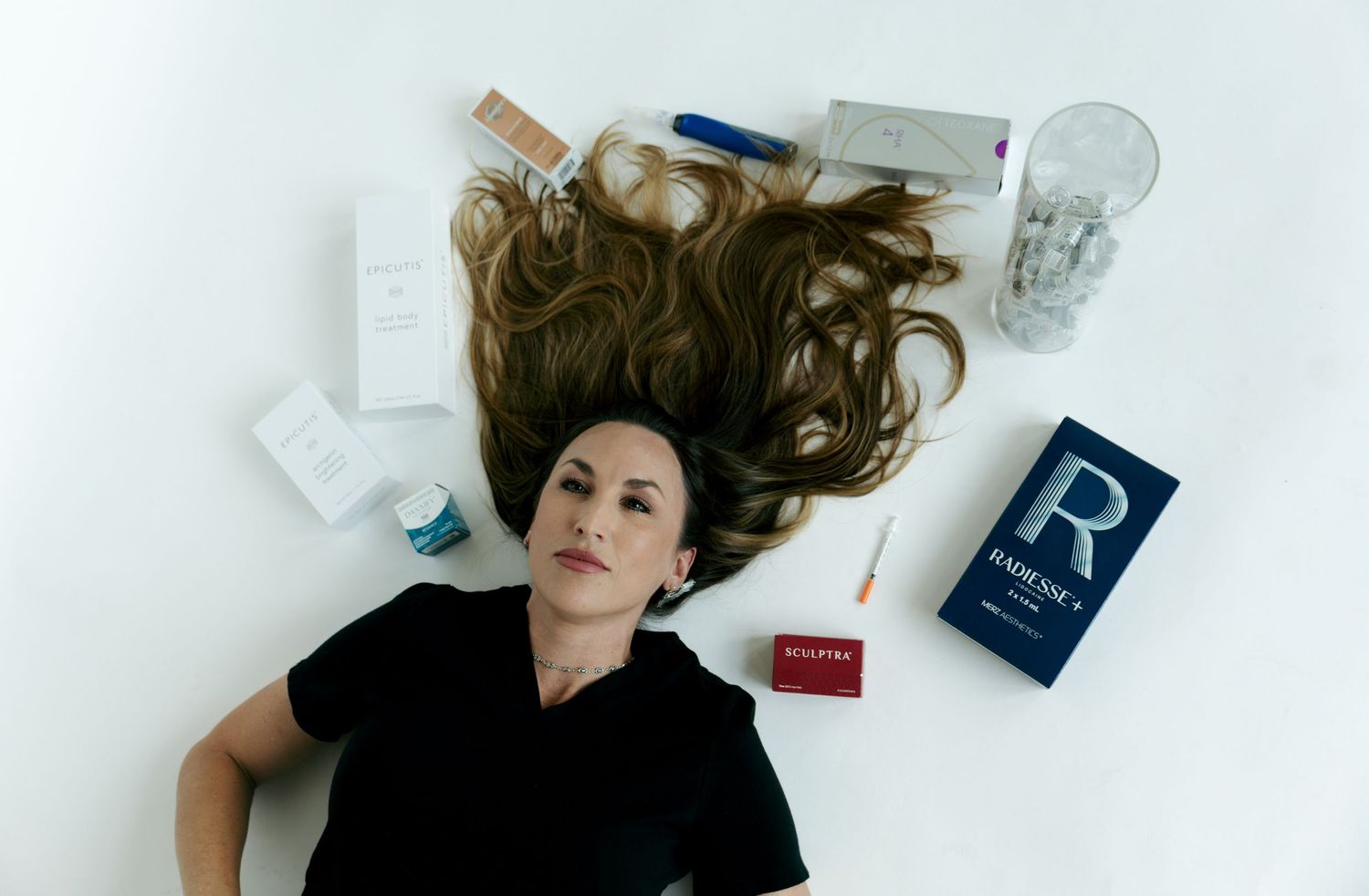 A woman in black lies on a white floor surrounded by skincare and medical aesthetic products.
