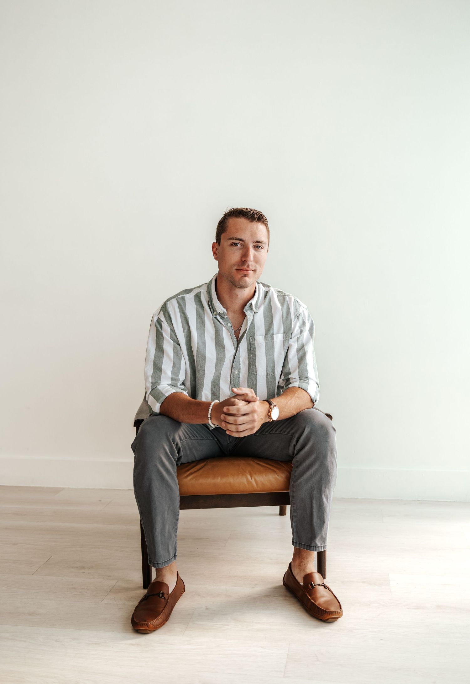 Young man in striped shirt and gray pants sitting in leather chair against white wall.