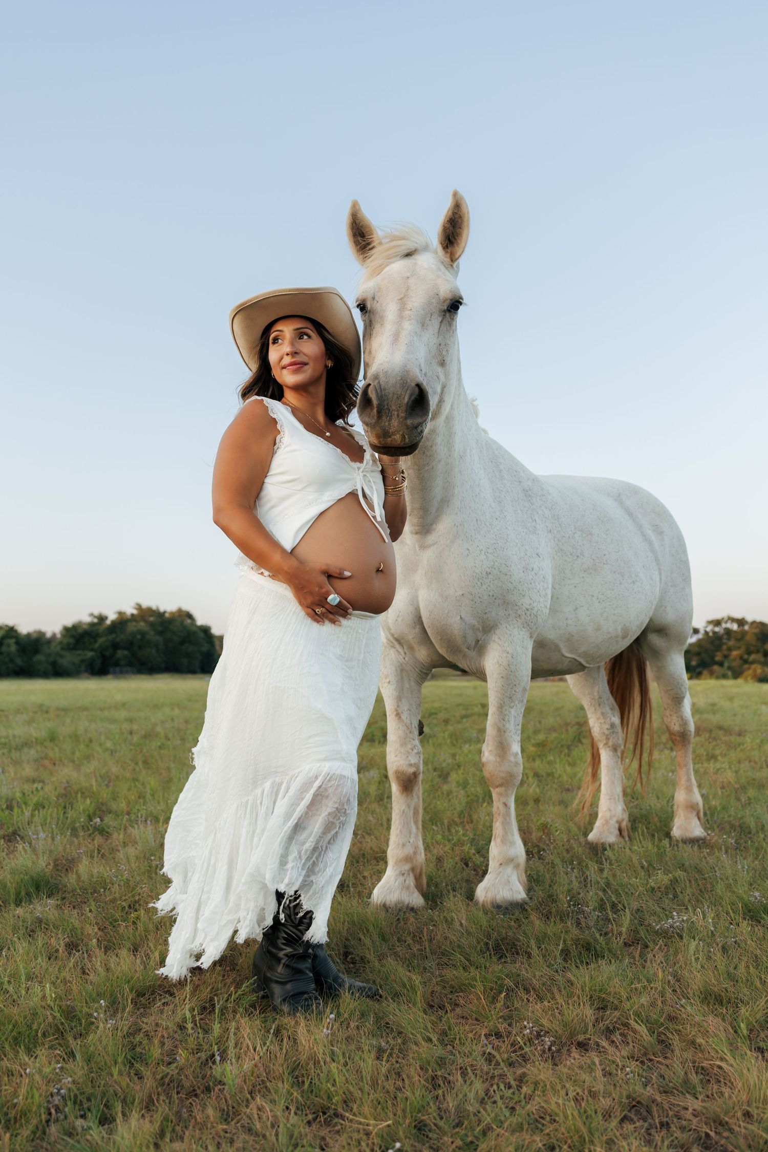 Pregnant woman in white dress and cowboy hat poses beside a white horse in a green field.
