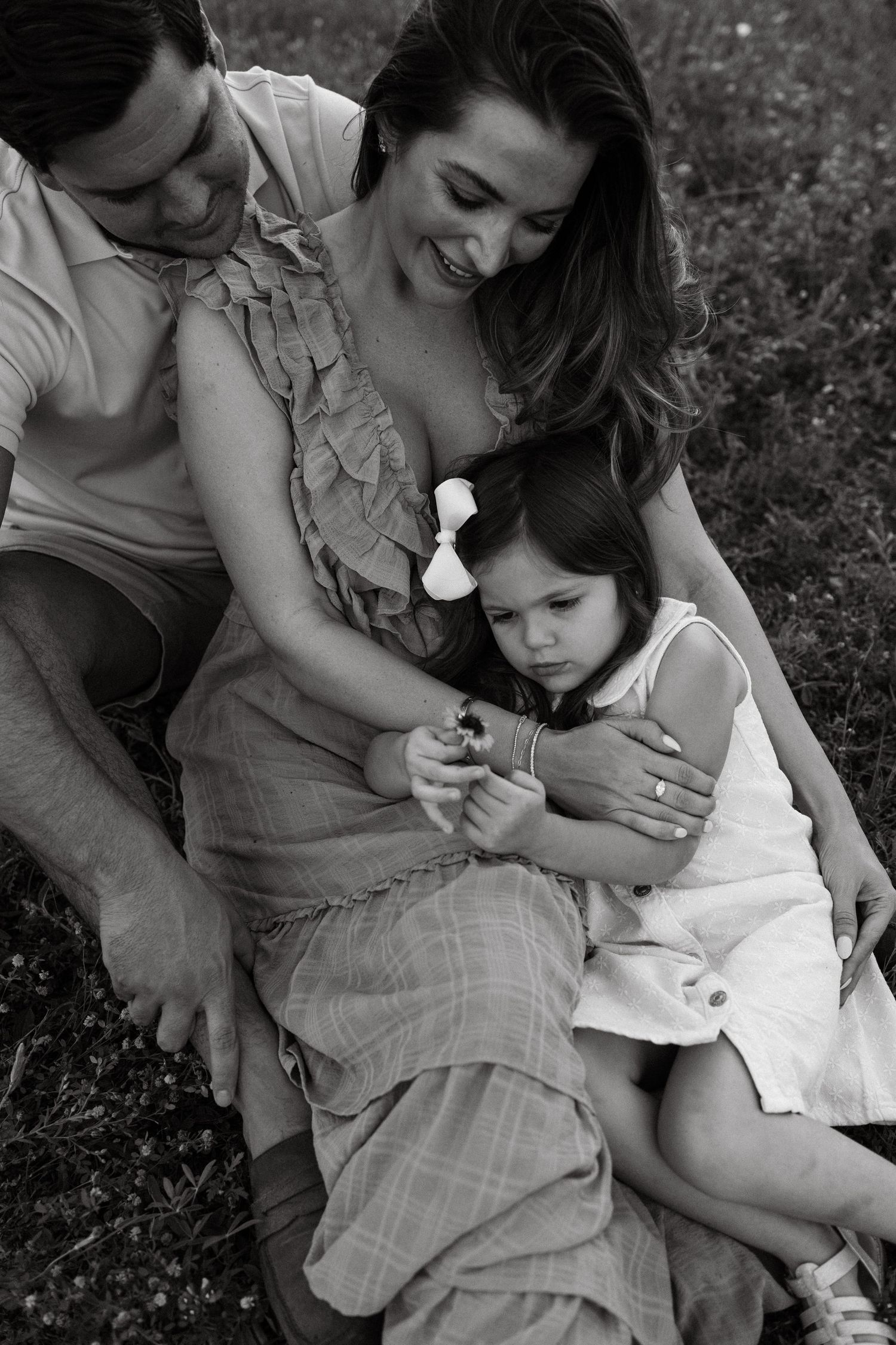 Black and white photo of parents smiling as their young daughter sits between them on the grass.