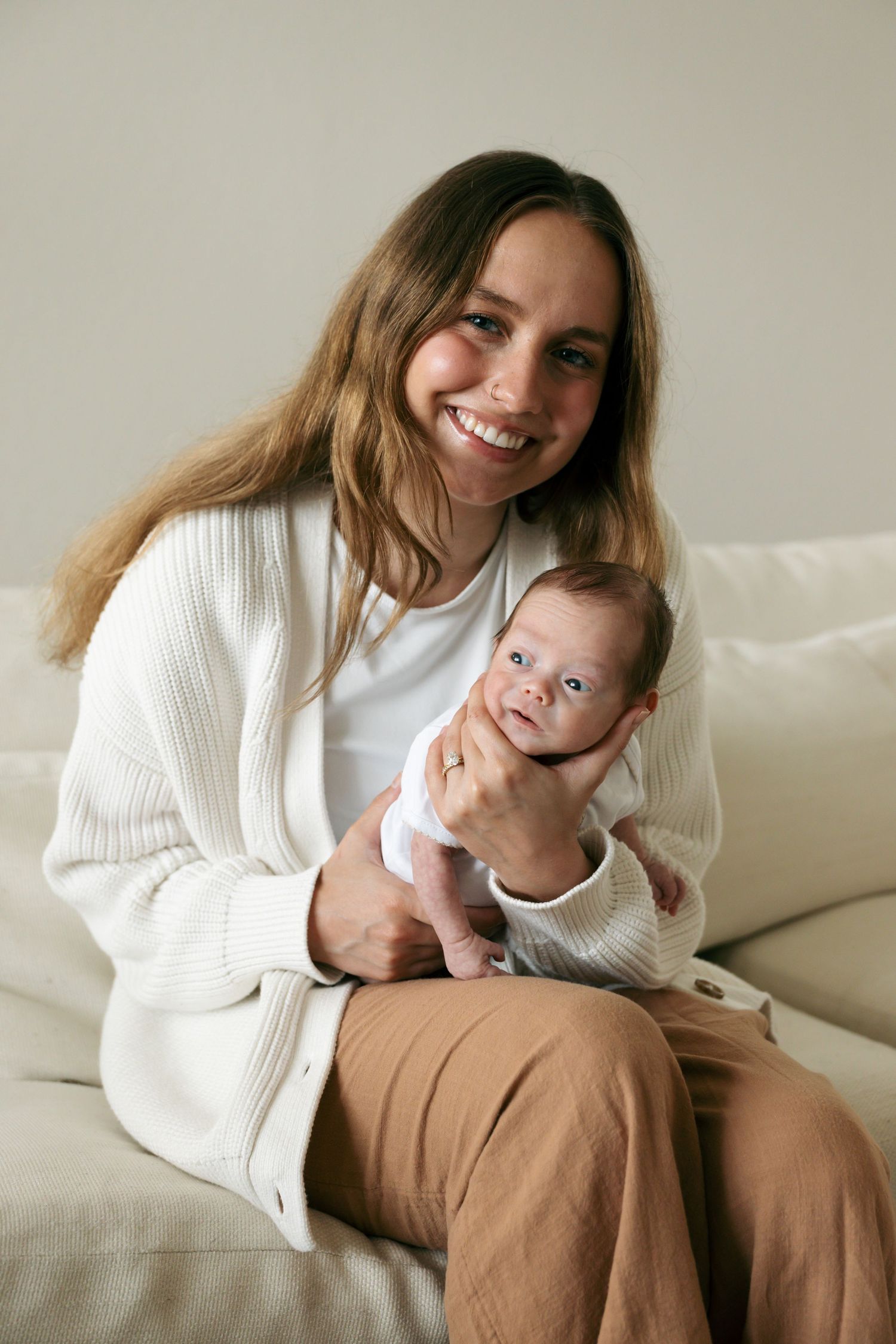 Smiling young mother in white cardigan holds newborn baby while sitting on cream couch indoors.