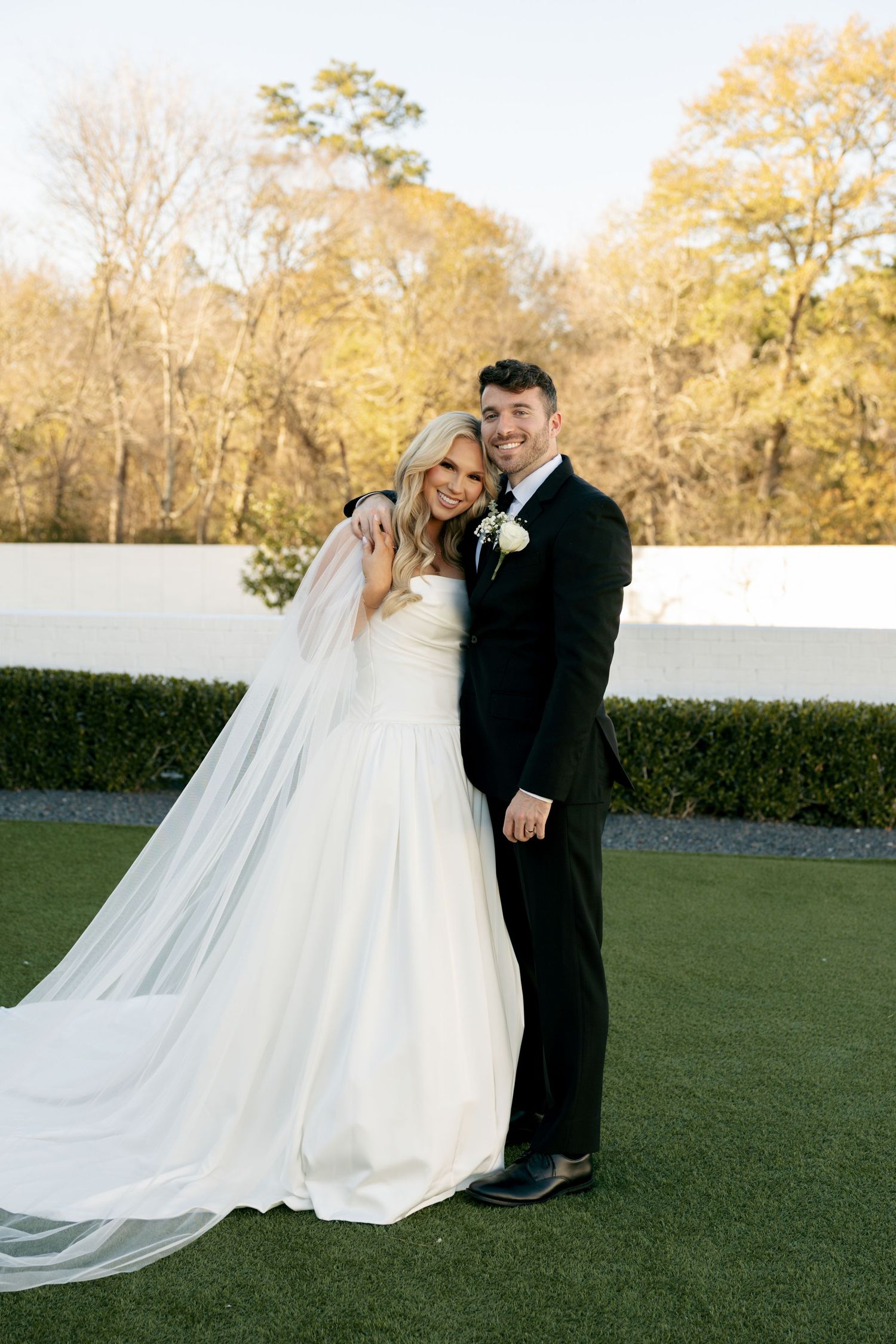 Smiling bride in strapless white gown with flowing veil and groom in black suit pose outdoors on green lawn.