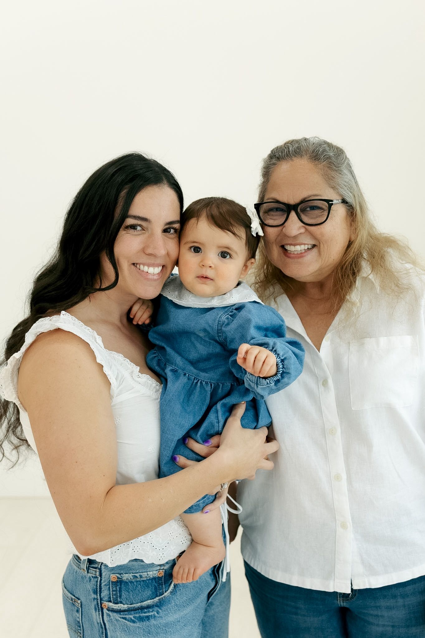 Three generations of women smiling together, with grandmother, mother holding baby in blue dress.